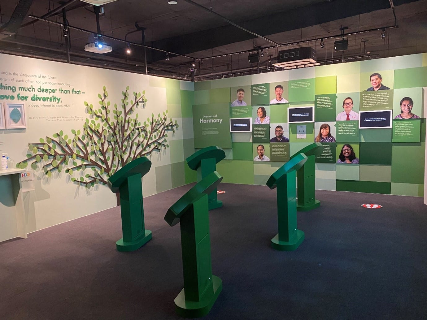 A museum exhibit room with green kiosks, a decorative tree, and a wall of portraits titled Humans of Harmony.