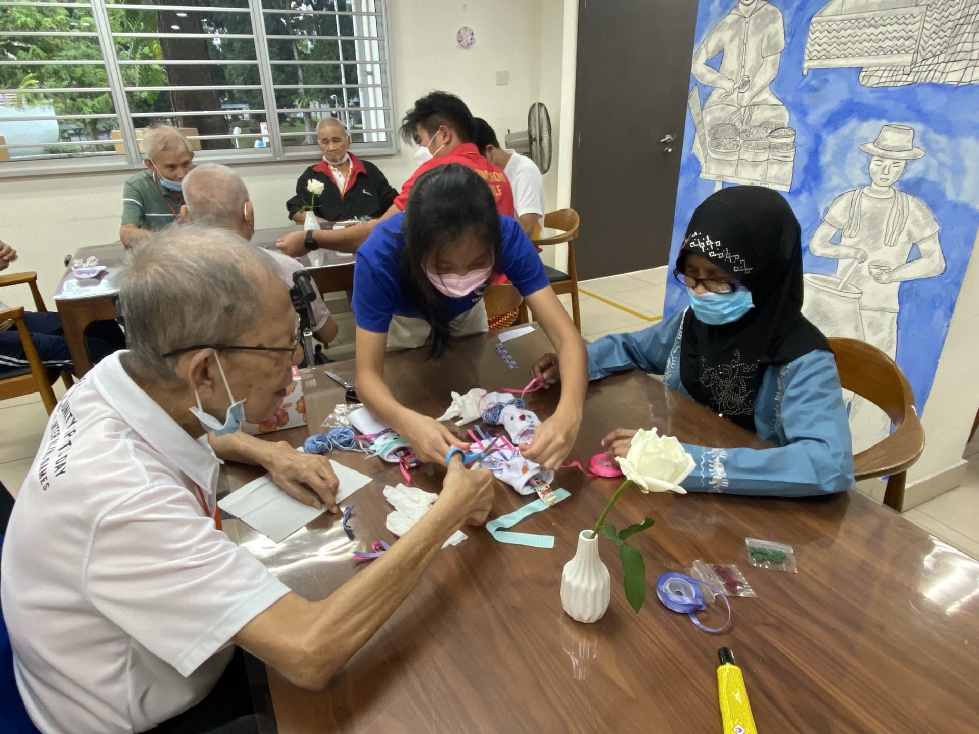 Elderly people and masked volunteers doing a craft activity with ribbons at a table, with a white flower in a vase nearby.