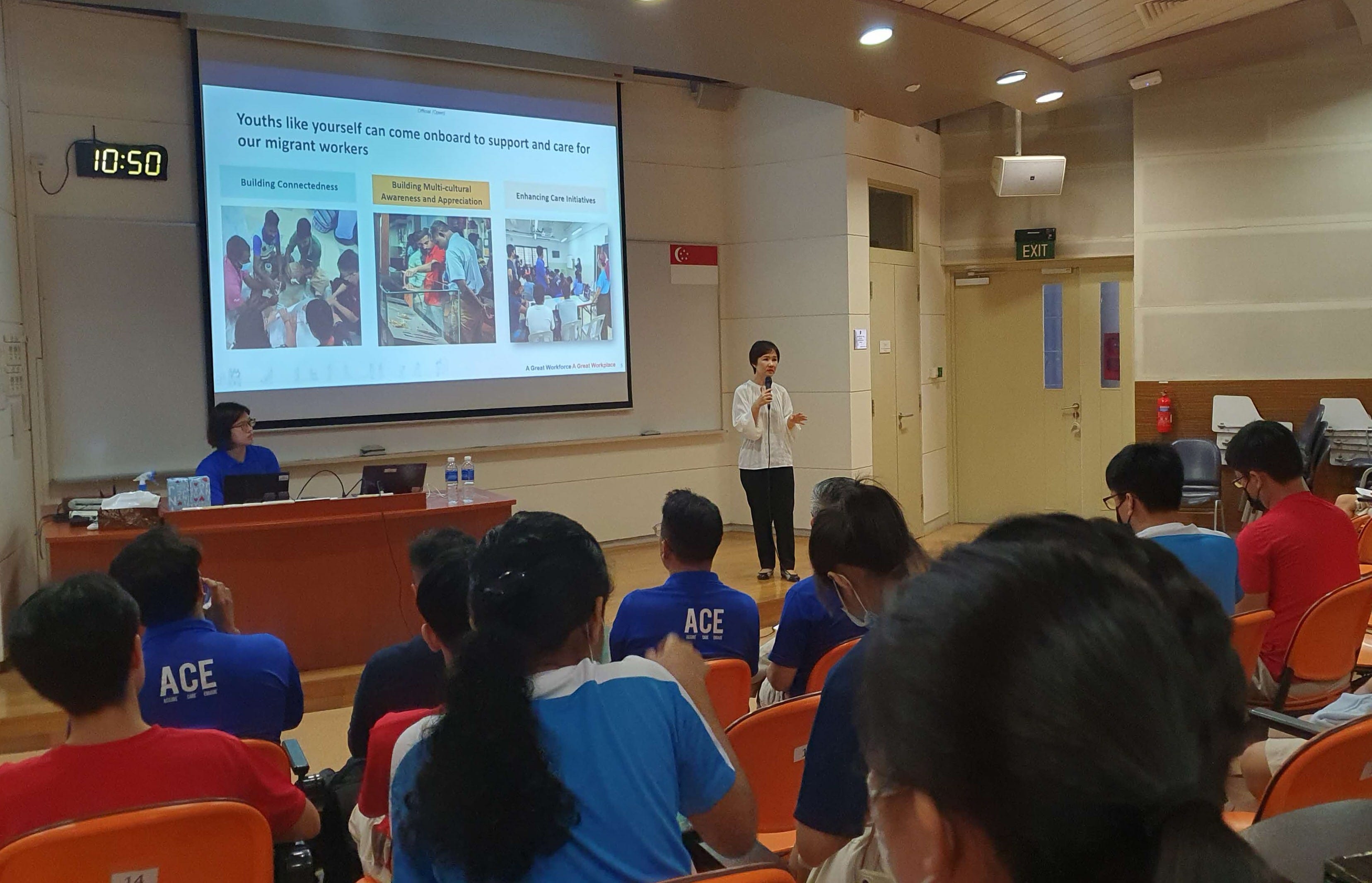 A woman speaks at a lecture hall about supporting migrant workers, with an audience in blue ACE shirts seated in rows.