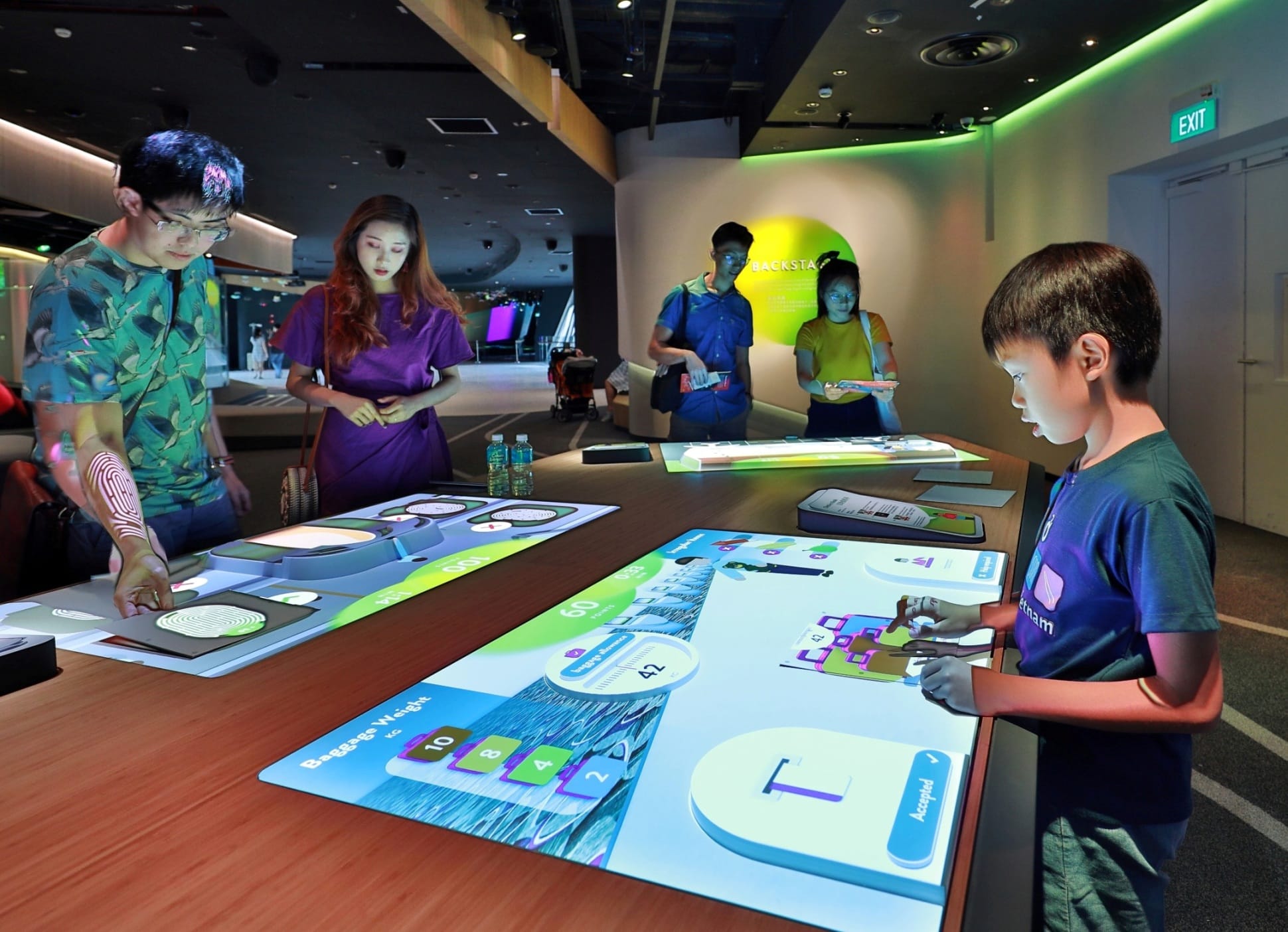 Several people including a young boy interact with large illuminated touch-screen tables displaying colorful interactive games in a museum exhibit.