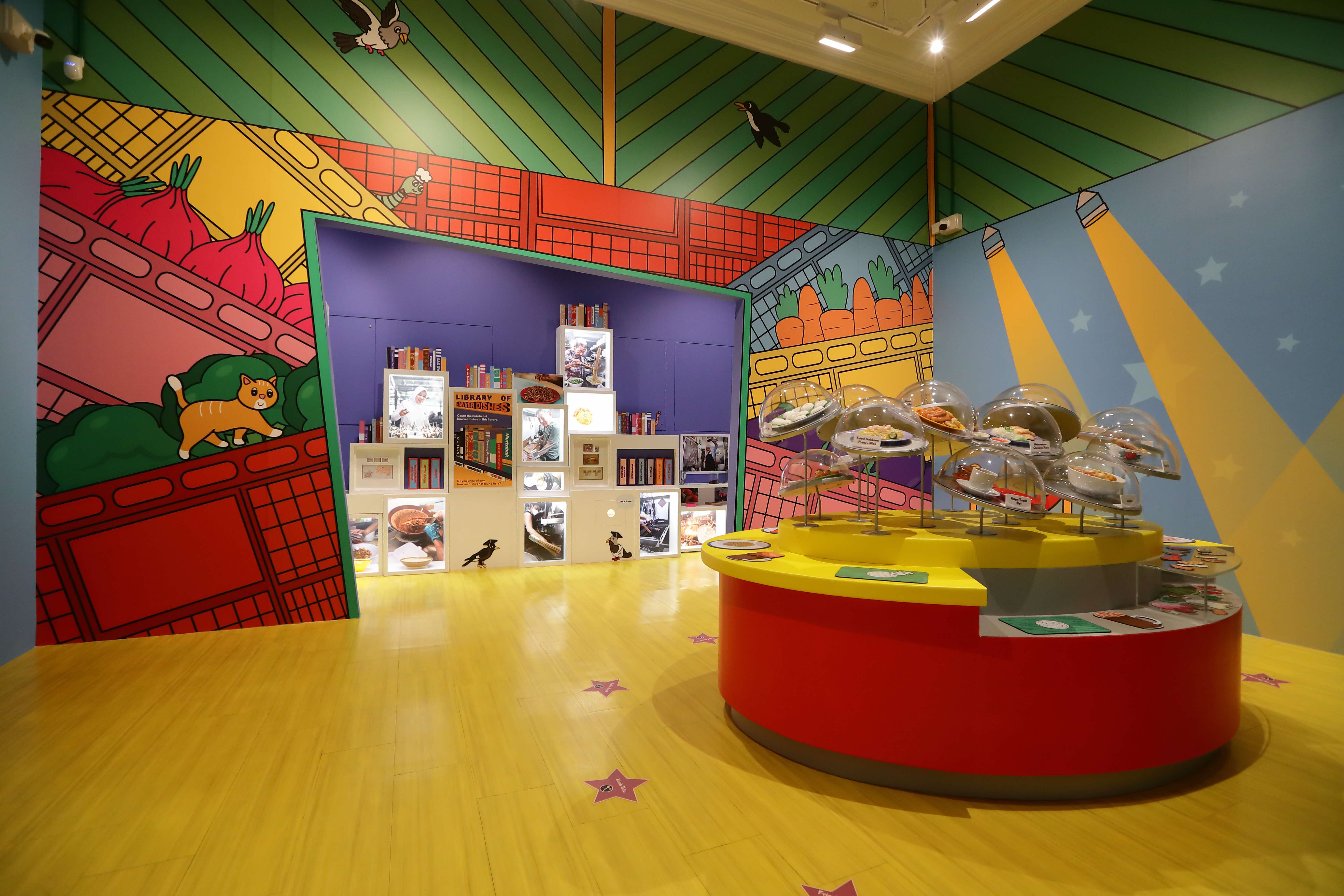 Colorful museum exhibit room with bold cartoon murals, a round yellow display stand with domed food models, and a photo display wall.