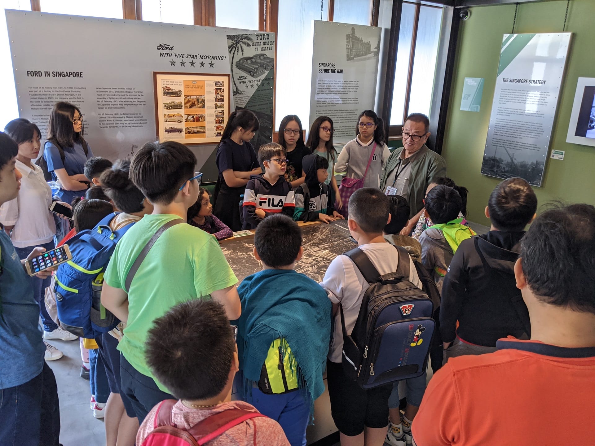 A group of children with backpacks gather around a display table in a museum with Singapore history exhibits on the walls.