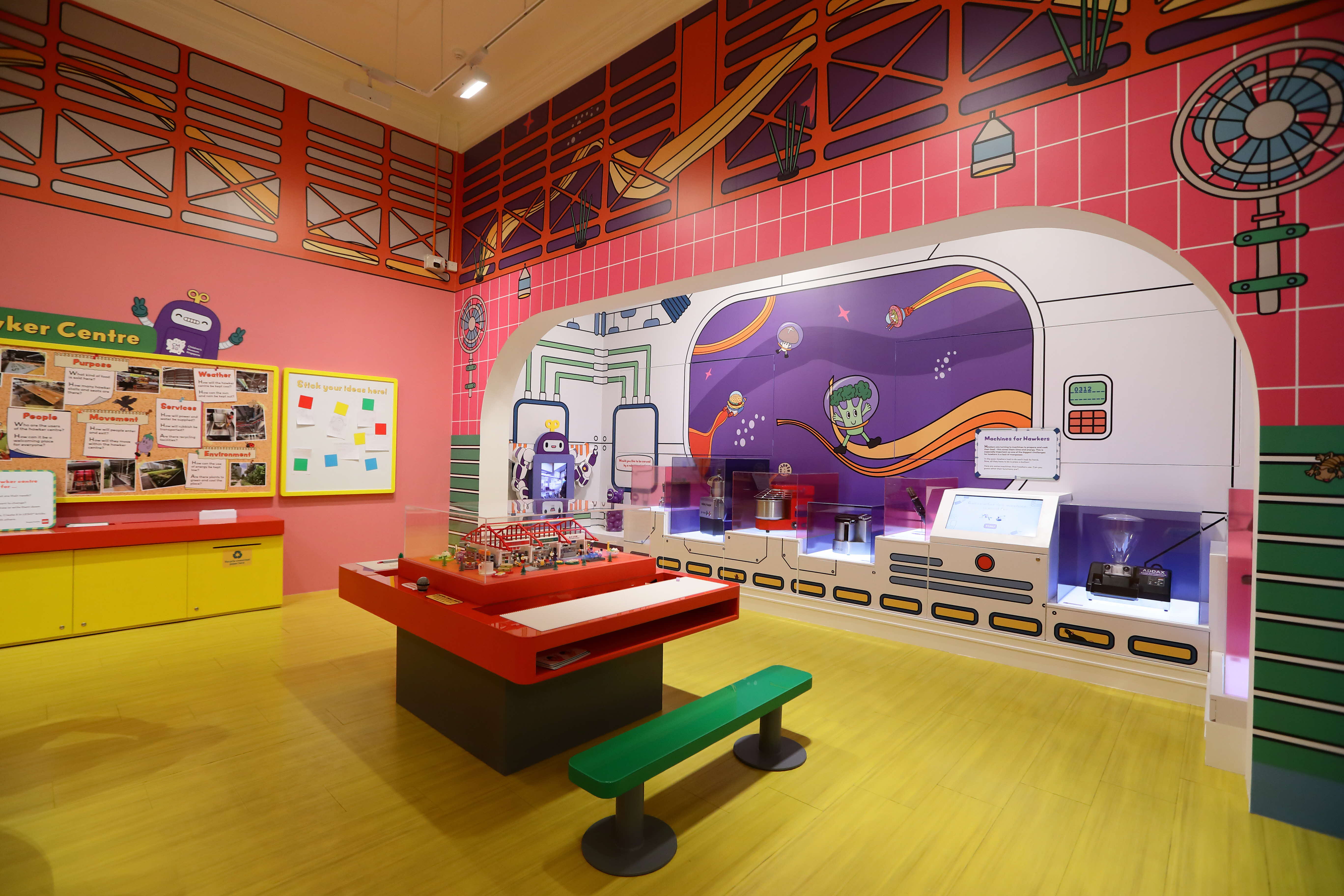 Colorful children's museum exhibit room with bright murals, interactive displays, a red table, green bench, and bulletin boards.