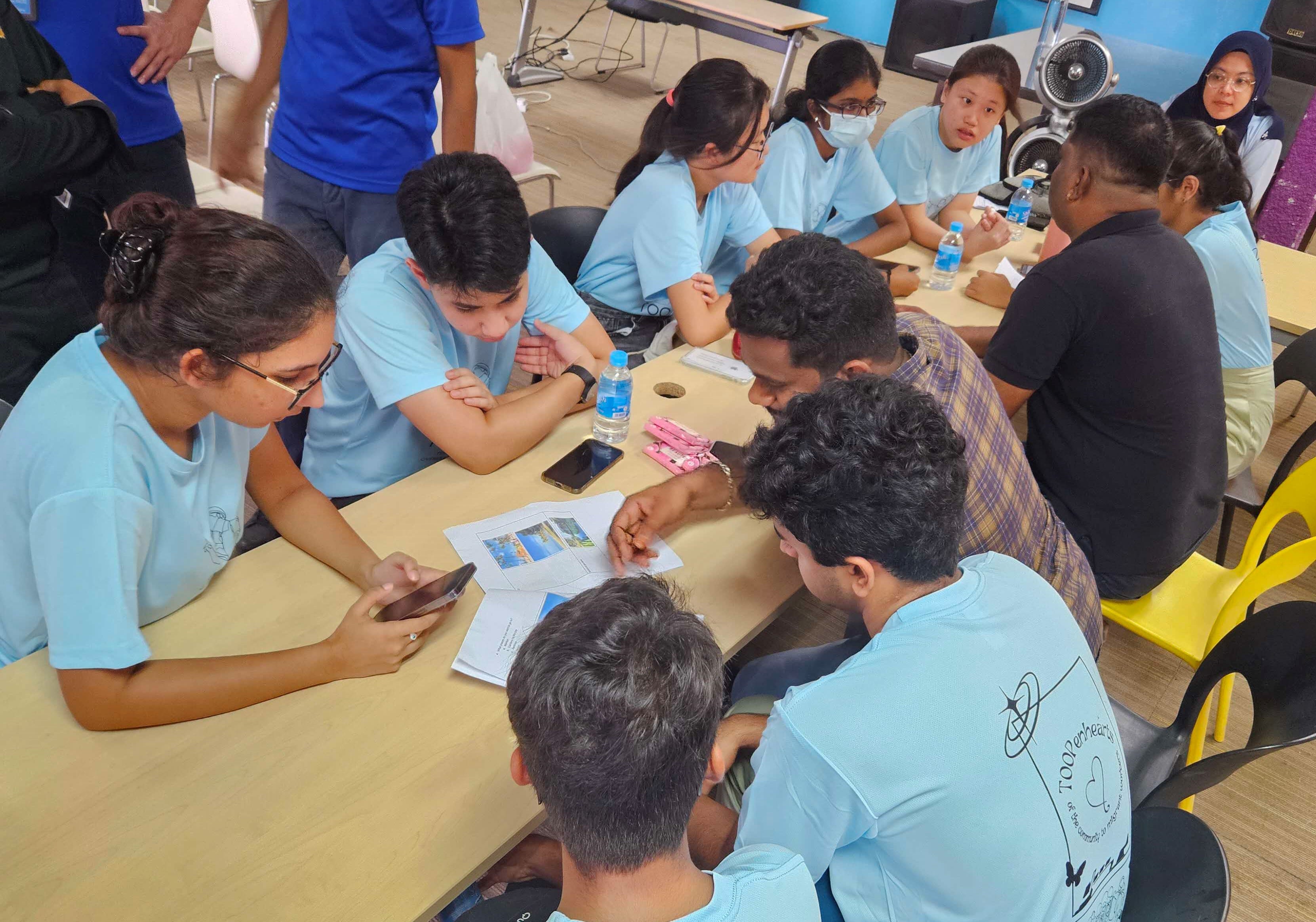 Students in light blue shirts gather around tables, reviewing papers and phones during a group activity indoors.