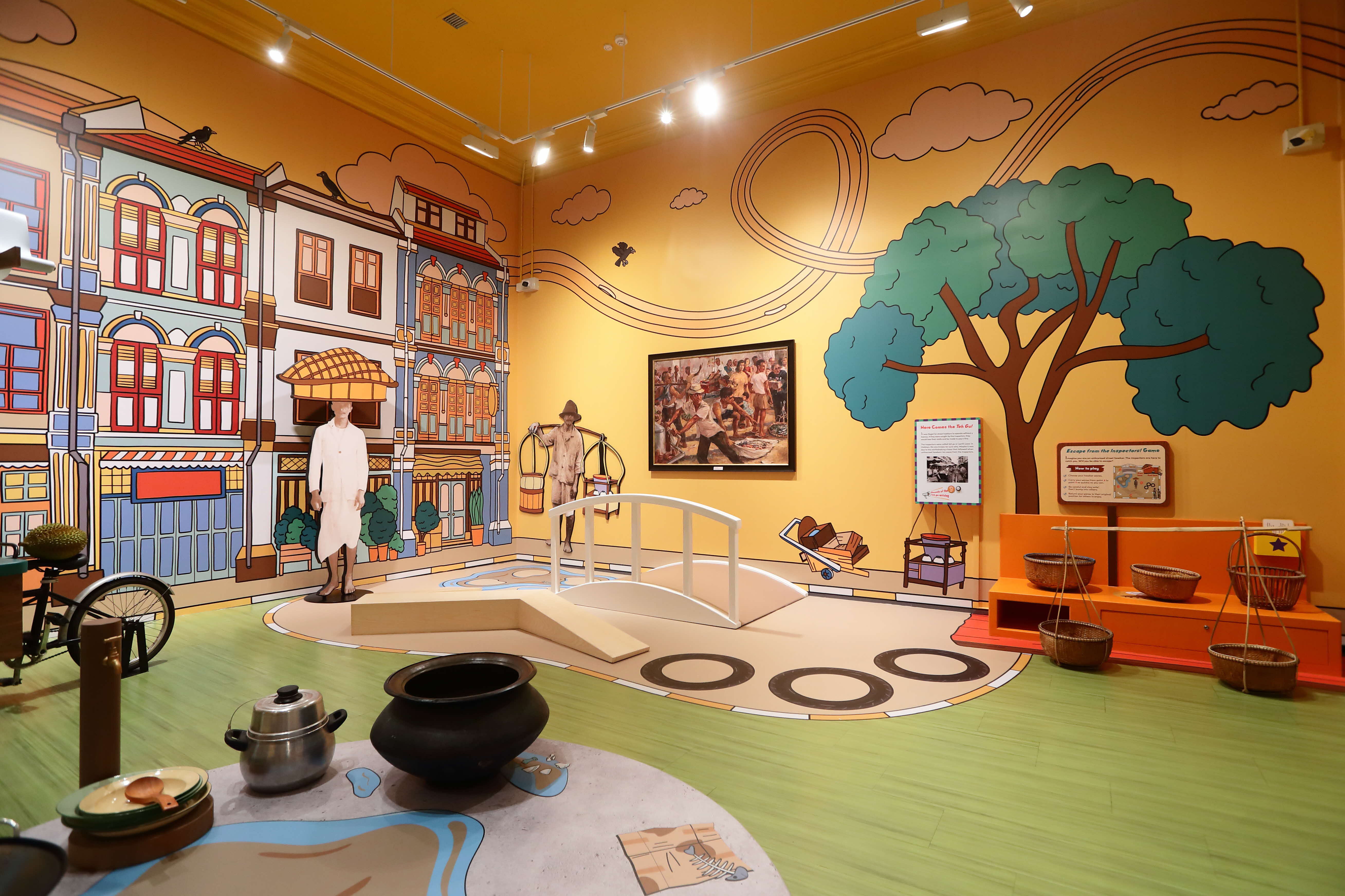 Colorful museum exhibit with cartoon murals of shophouses, mannequins, baskets, pots, and a small bridge on a green floor.