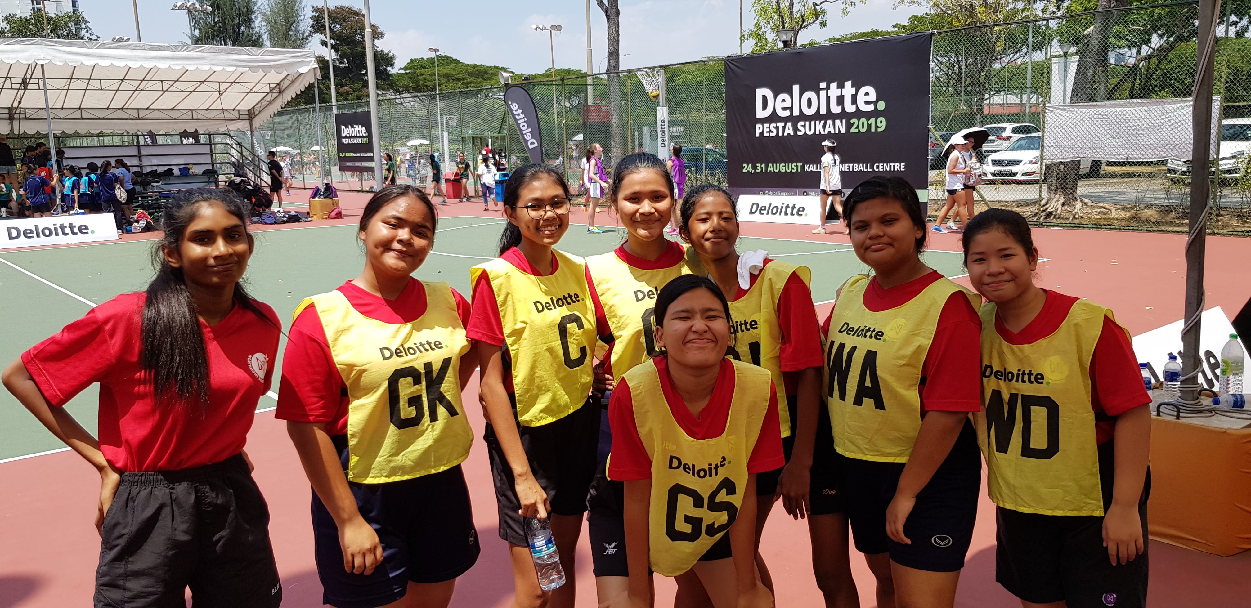 Eight young girls in red shirts and yellow Deloitte netball bibs pose on a court at Deloitte Pesta Sukan 2019.