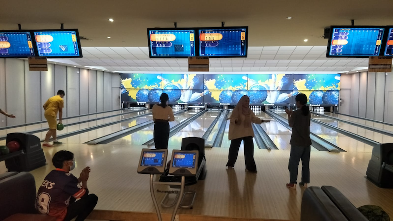 Several people bowling at a modern indoor alley with scoring screens overhead and a colorful video wall at the end.