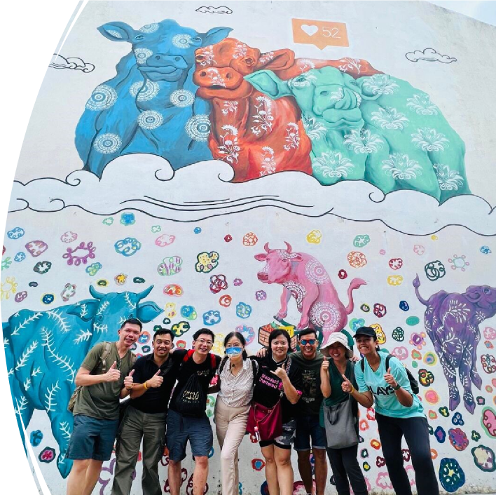 Eight people pose with thumbs up in front of a colorful mural featuring decorated cows in blue, red, and teal.