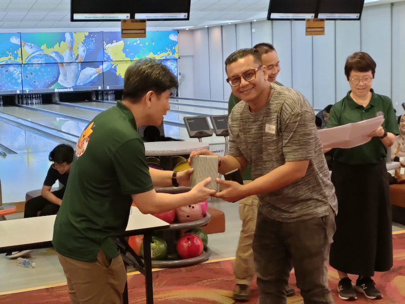 Two men exchange a prize at a bowling alley event, with bowling balls and lanes visible in the background.