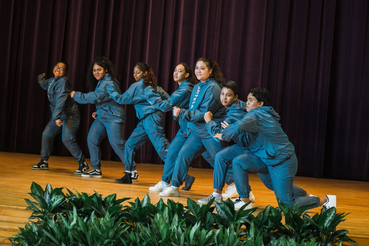 Seven young dancers in matching blue outfits perform a synchronized routine on a wooden stage with a red curtain backdrop.