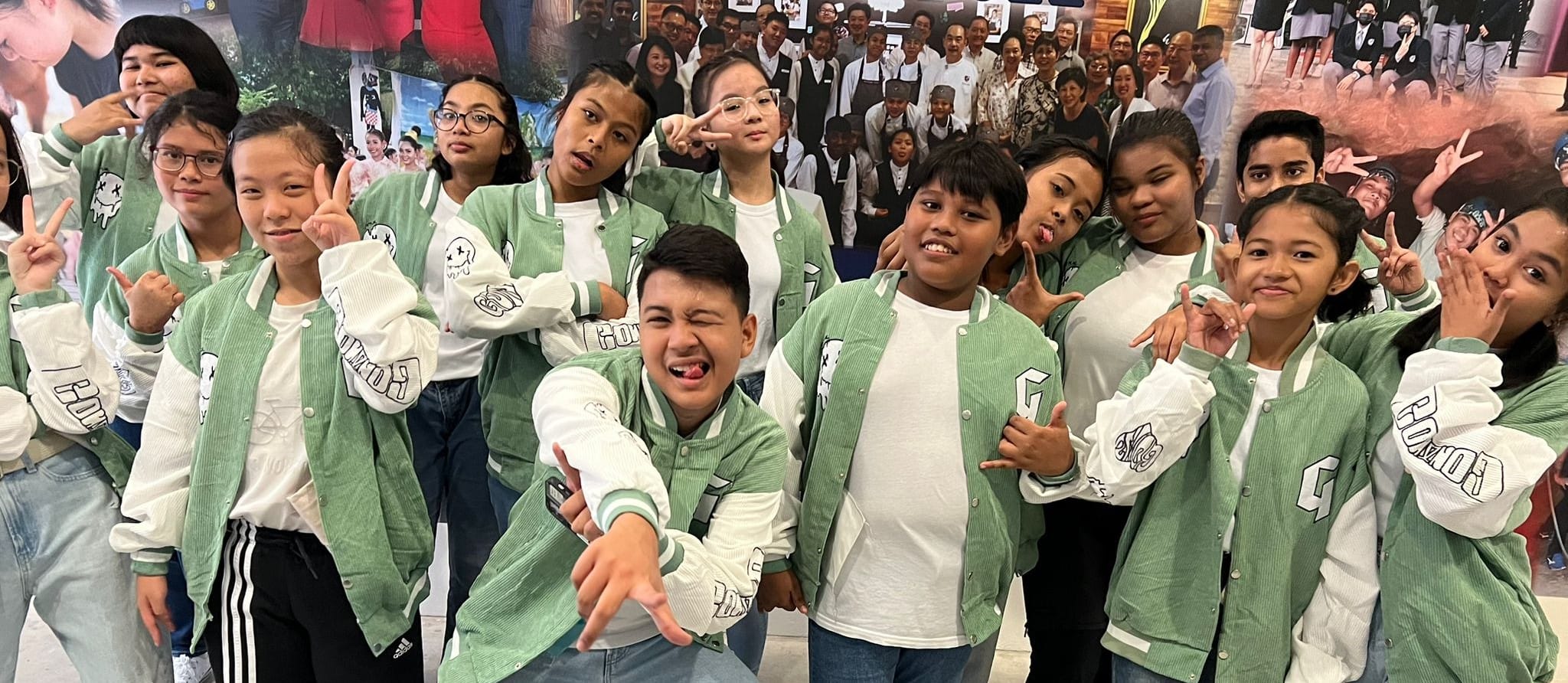 A group of children in matching green and white varsity jackets posing and making fun expressions together.
