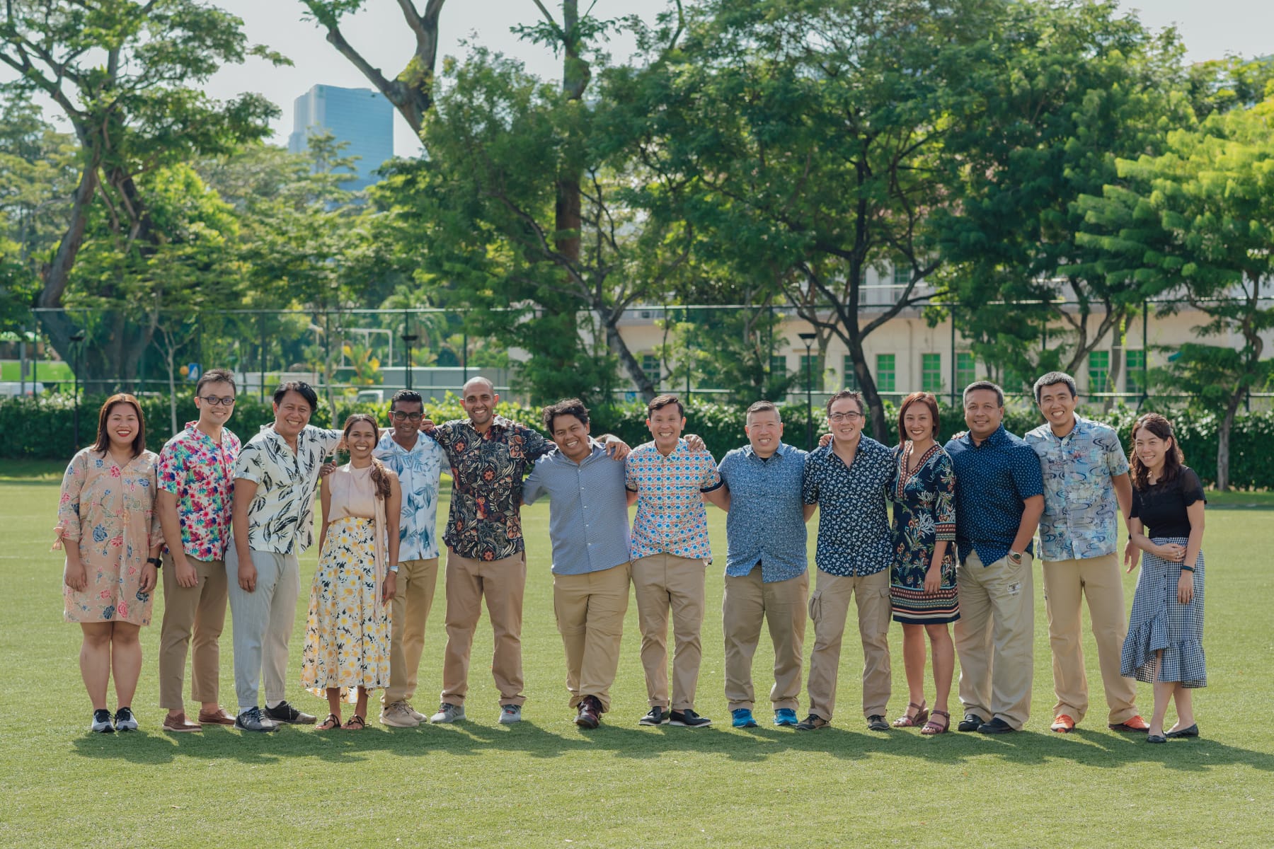 A group of about 14 smiling adults standing together on a grassy field with trees and a building behind them.