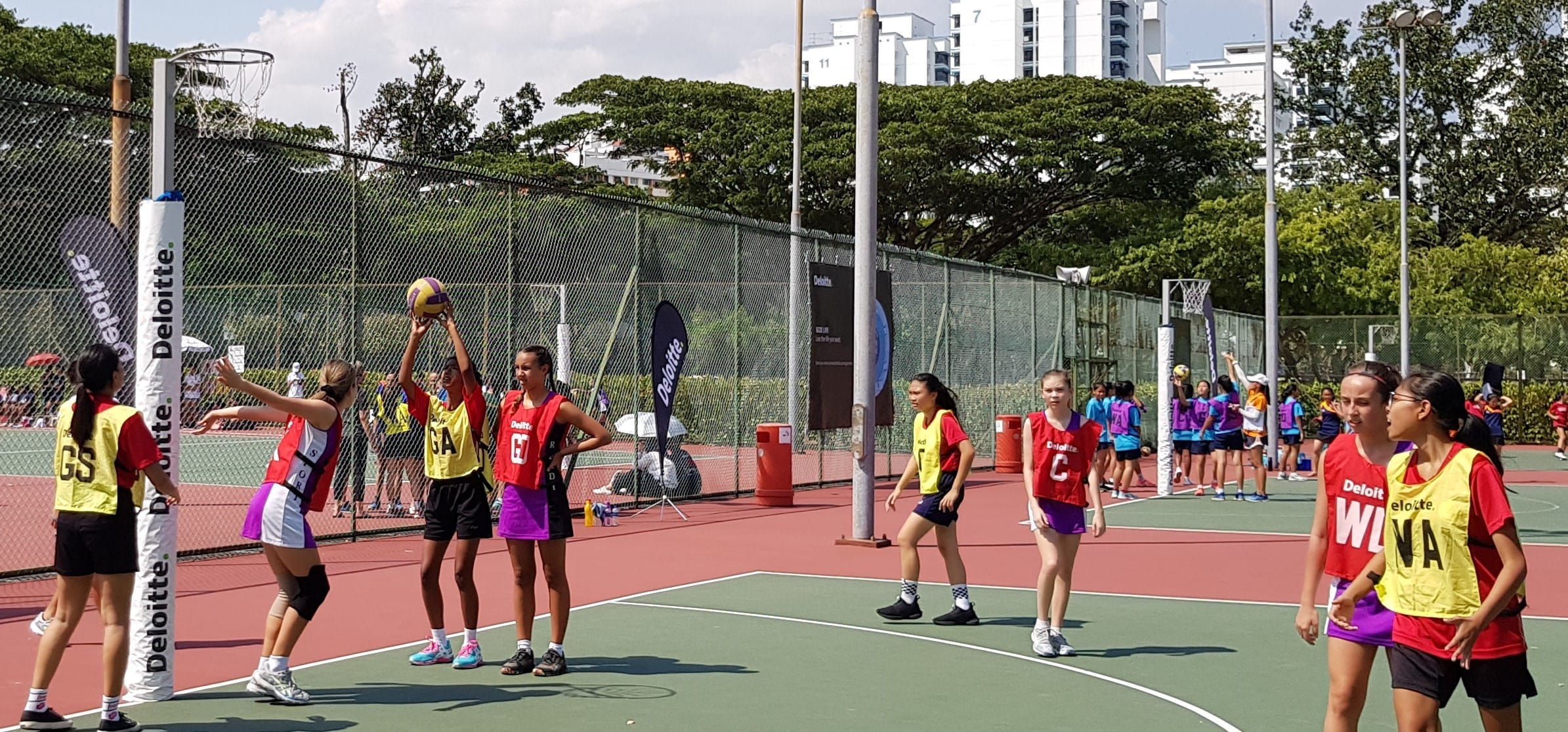 Girls playing netball outdoors, one player shooting toward the post while others defend on a red and green court.