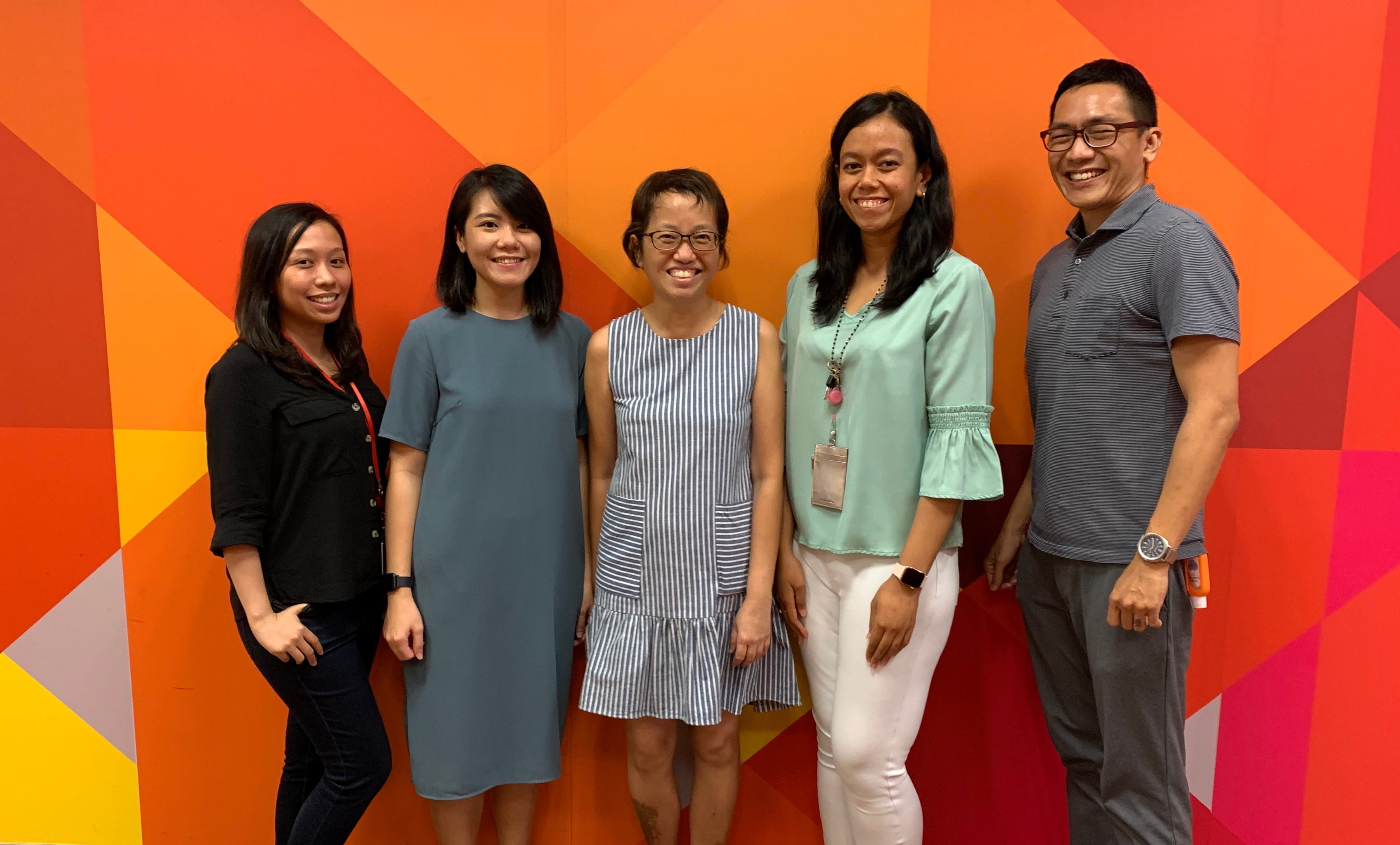 Five smiling people stand together in front of a bright orange geometric patterned wall.