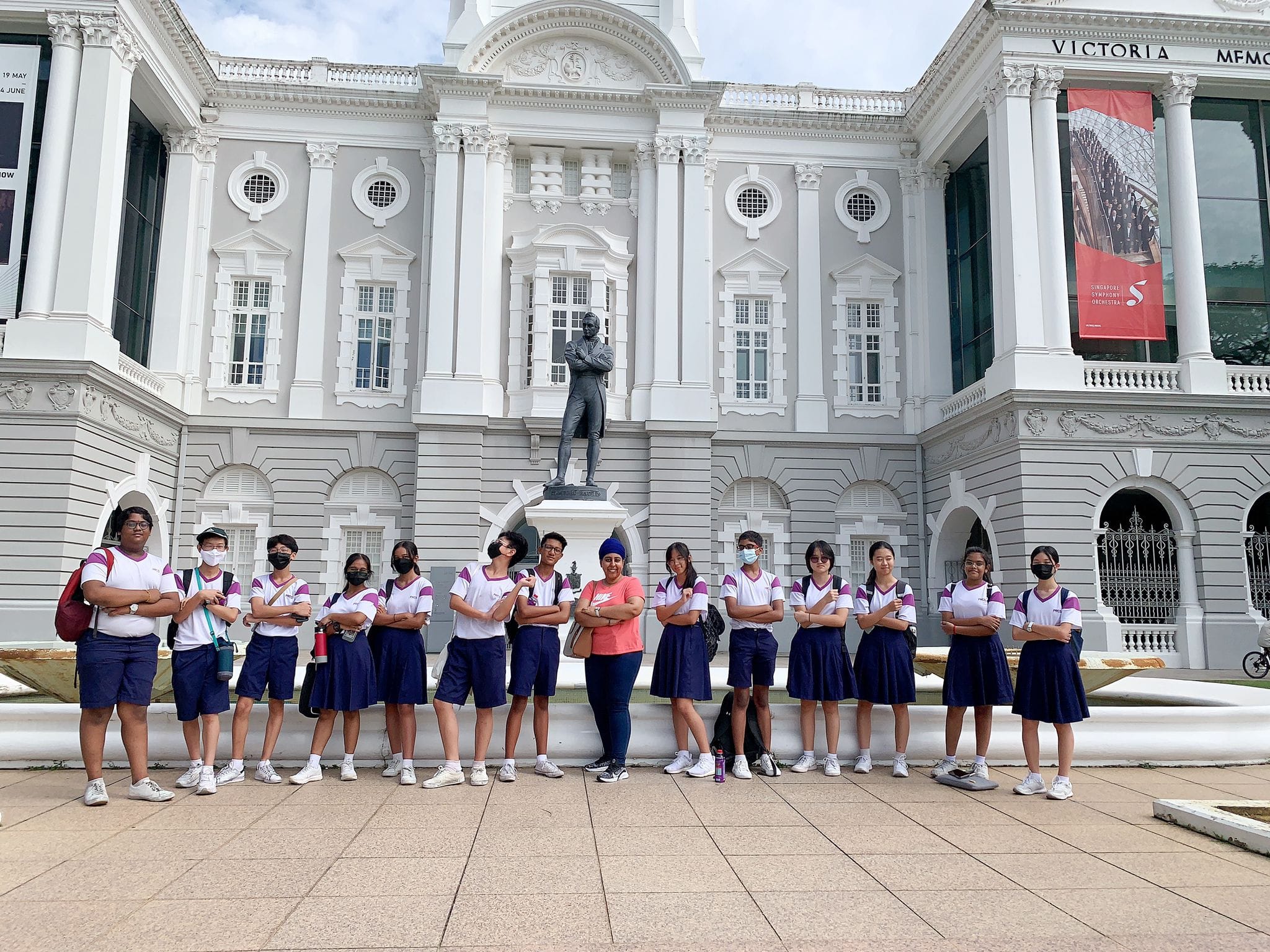 Students in school uniforms pose with arms crossed before Victoria Memorial Hall and a bronze statue.
