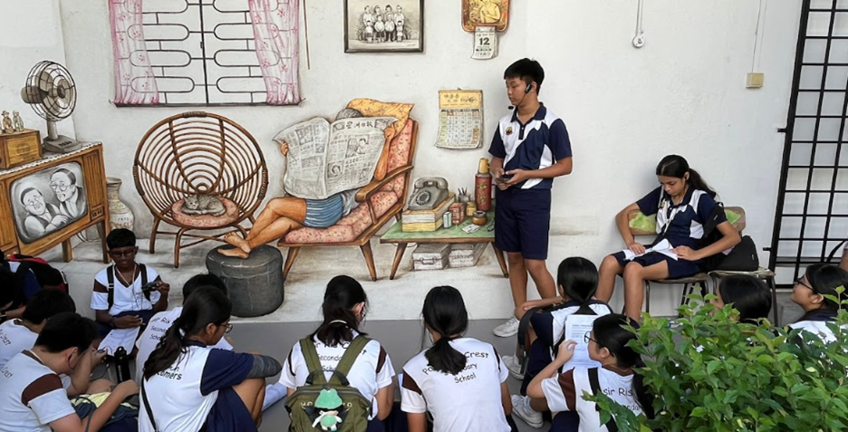 Students in school uniforms sit on the ground facing a boy presenting in front of a retro-themed wall mural.