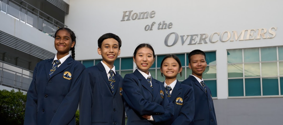 Five students in navy blazers and ties smile in front of a building reading "Home of the Overcomers."