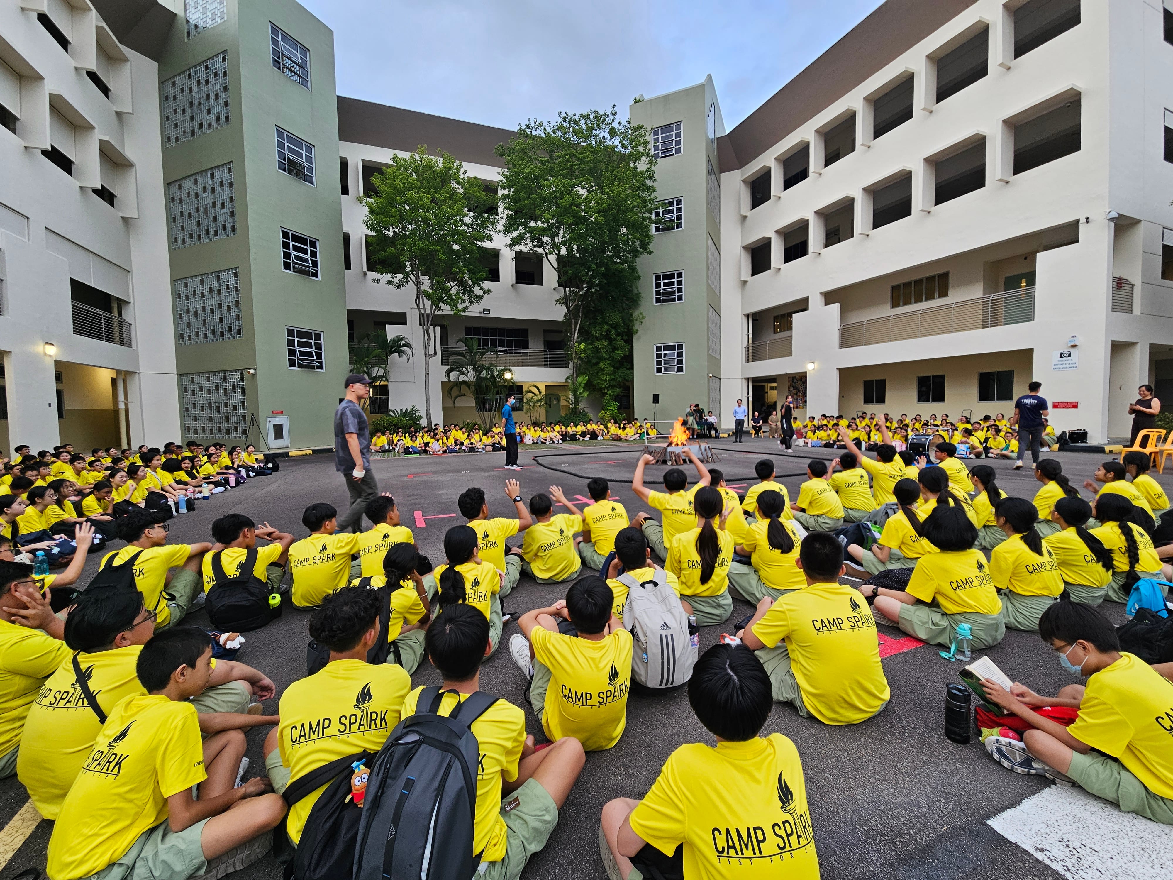 Photograph of a large outdoor gathering at a school courtyard showing students wearing matching yellow "Camp Spark" t-shirts seated on the ground facing a central stage area. The setting includes multi-story school buildings surrounding the courtyard, with several adults standing near the stage, indicating an organized camp event or assembly.