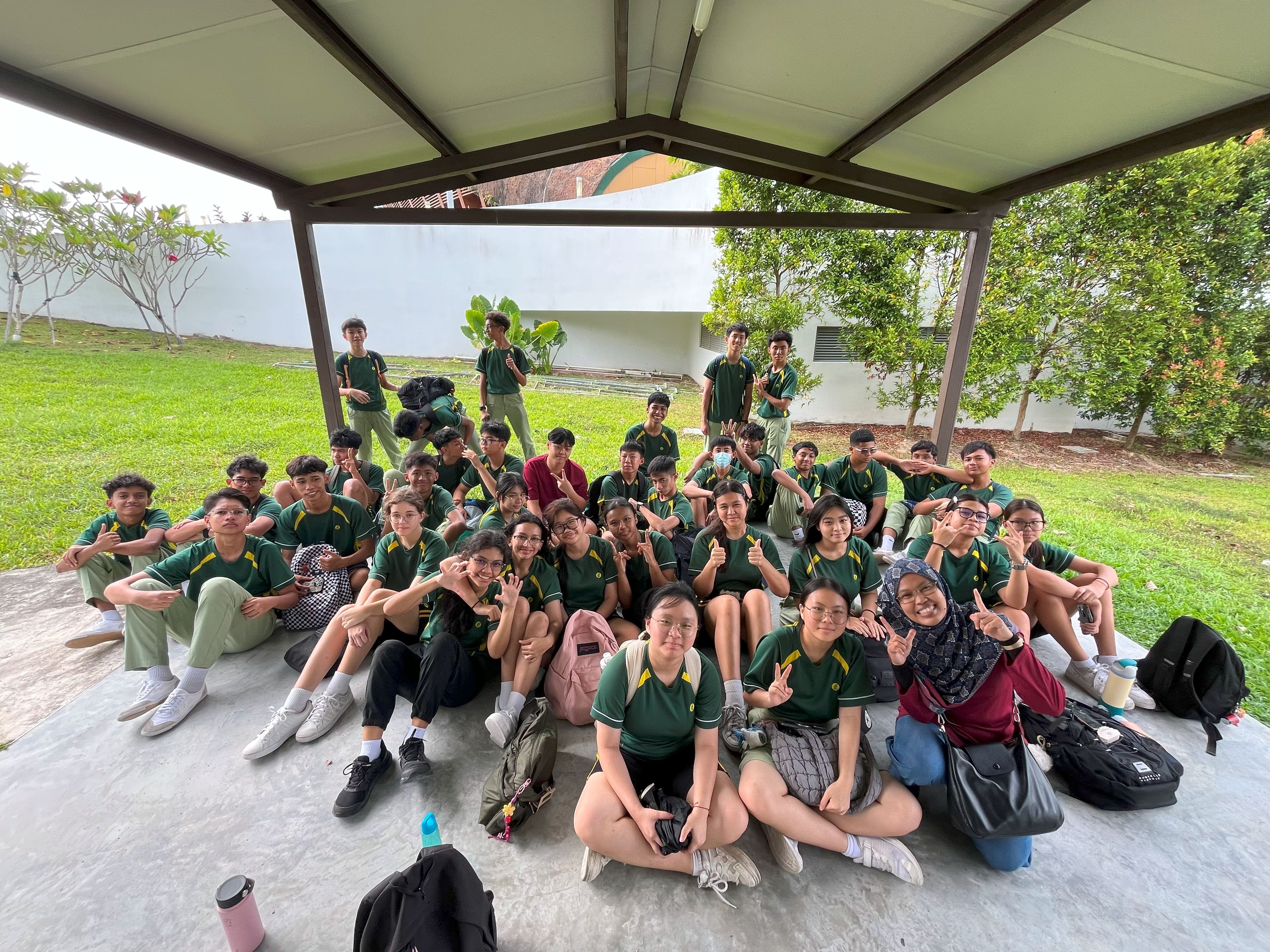 Photograph of a large group of students sitting and standing under a covered outdoor area, posing for a group photo. Most students wear matching green and yellow uniforms, with some making peace signs, and backpacks and water bottles are visible around them.