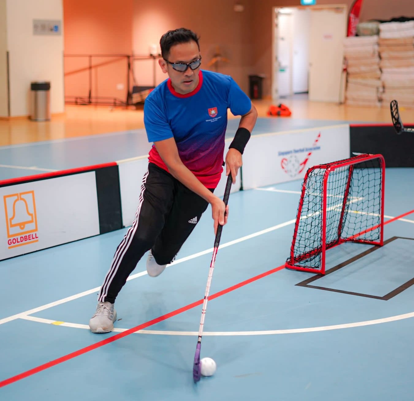 Man in blue and red jersey playing floorball indoors, holding a stick near a white ball with a red goal behind him.