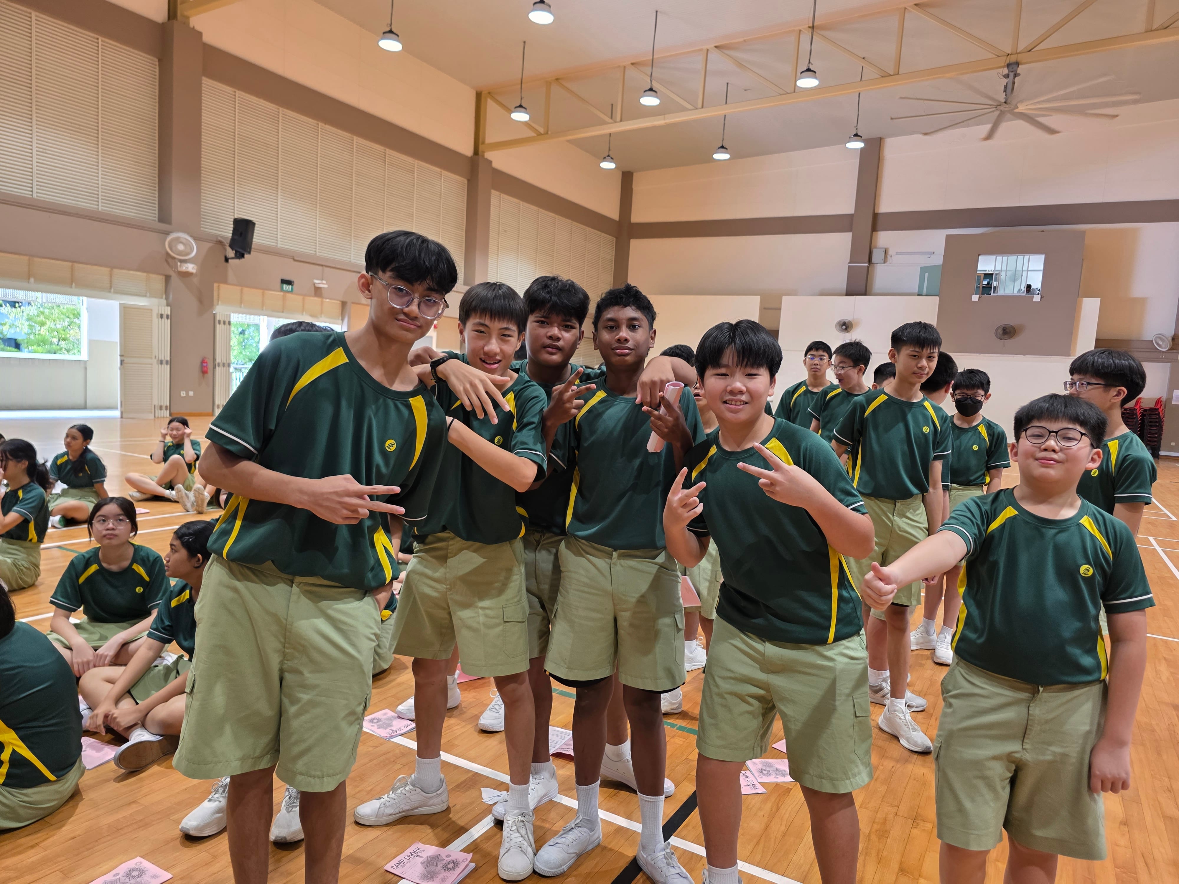 Photograph of a group of students in green and yellow sports uniforms posing and making peace signs inside a gymnasium. Other students in similar uniforms are seated or standing in the background, indicating a school event or sports activity.