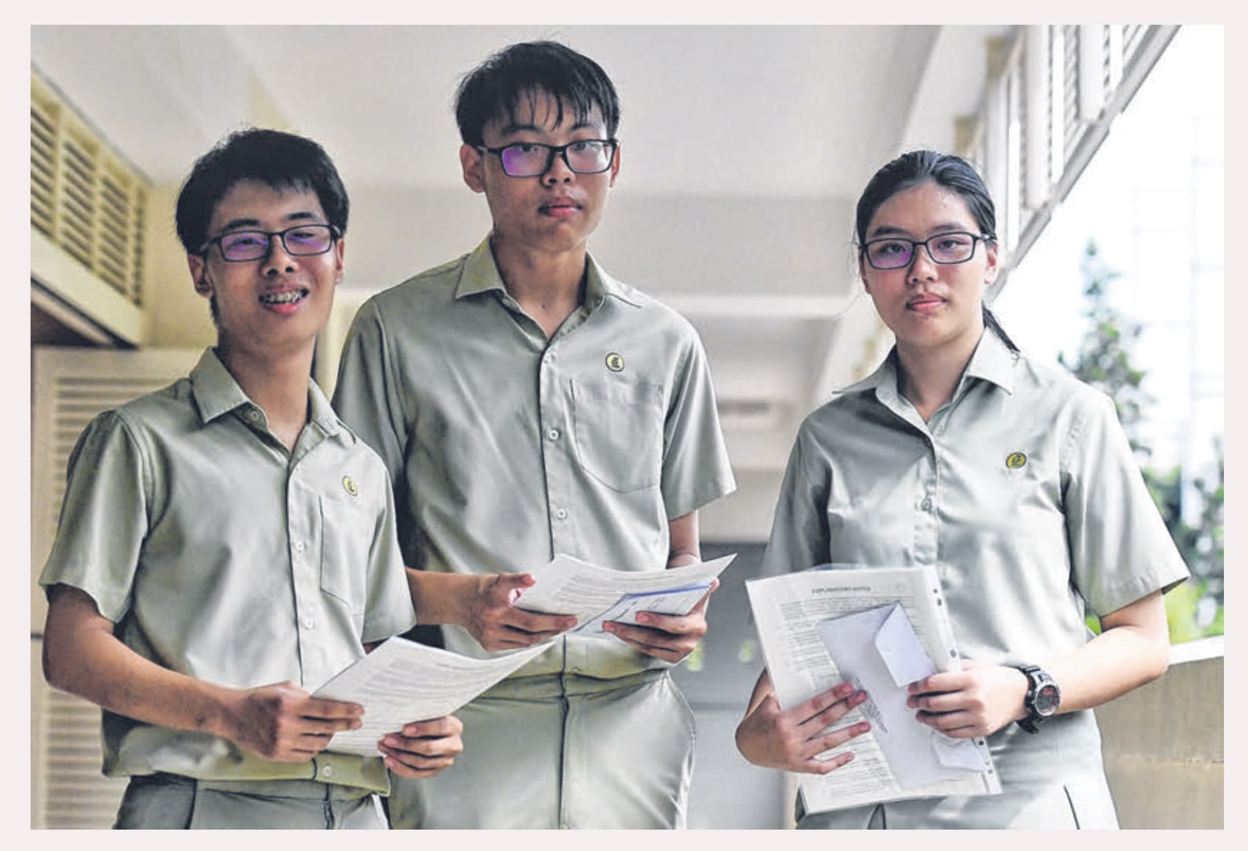 Three students in matching gray uniforms and glasses hold papers while standing in a school corridor.
