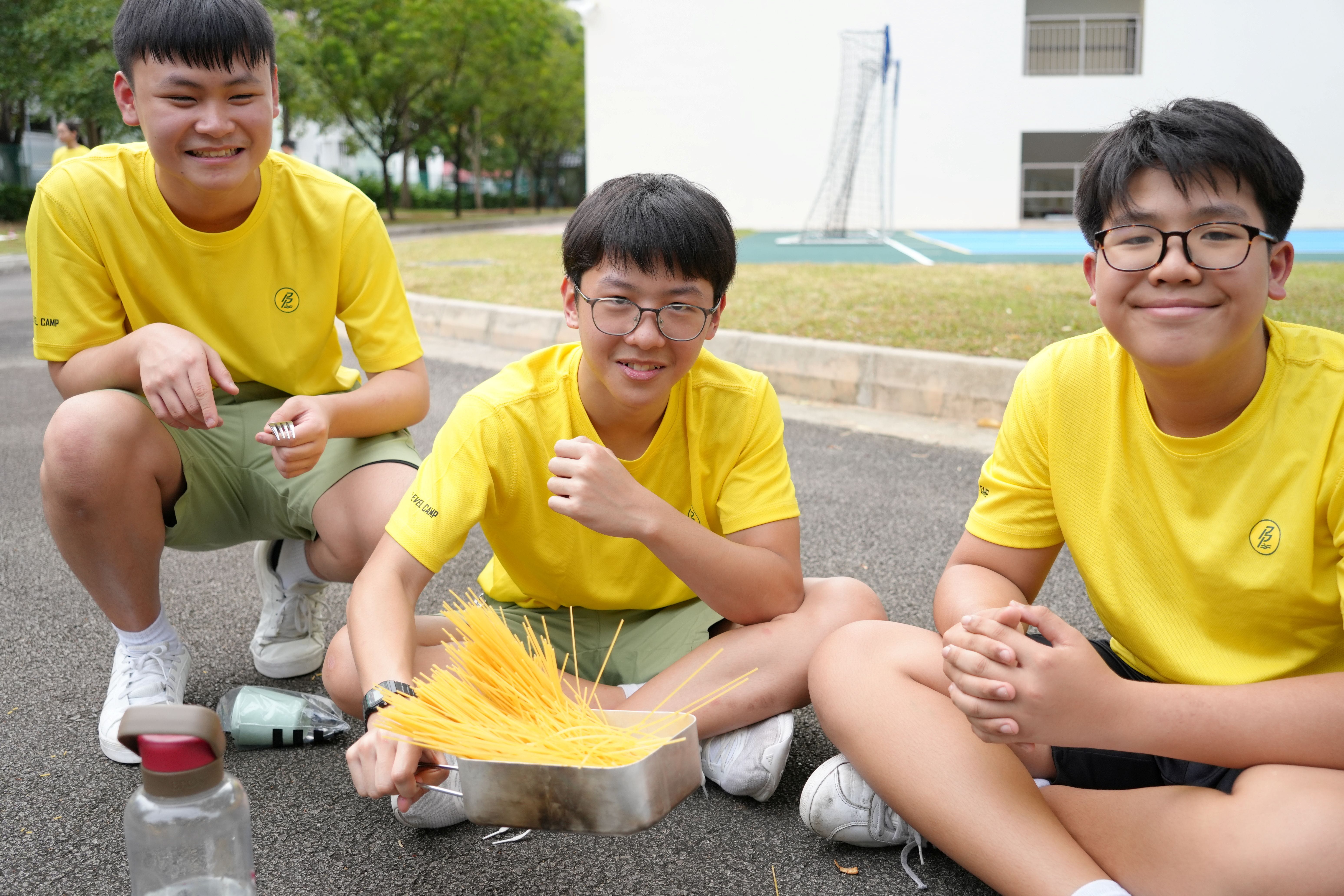Photograph of three boys wearing matching yellow shirts sitting outdoors on pavement near a sports field, engaging in a hands-on activity with uncooked spaghetti in a metal container. The scene highlights teamwork and experimentation, with a water bottle placed nearby and a building in the background.