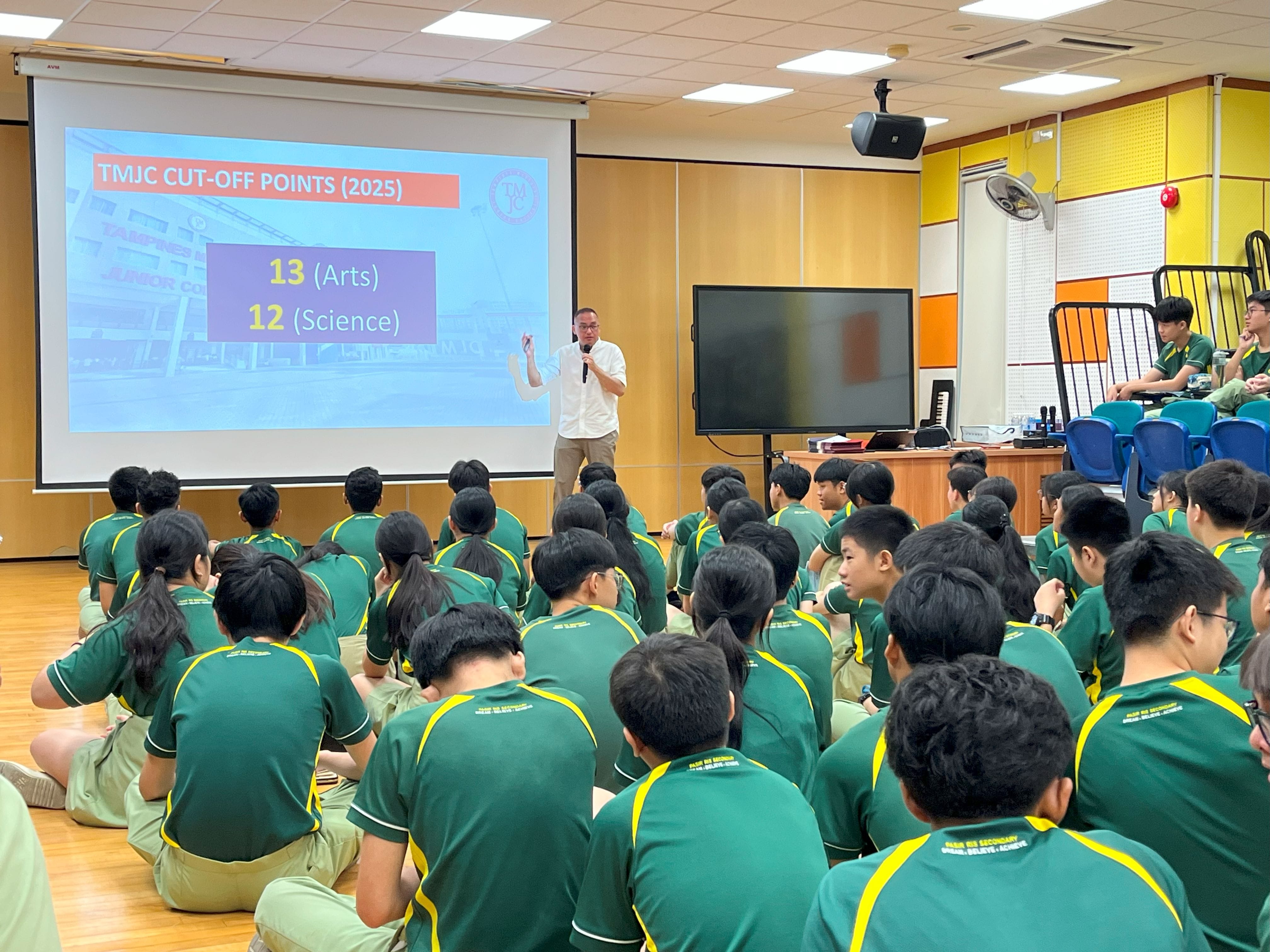 Photograph of a classroom presentation showing TMJC cut-off points for 2025, with a slide displaying "13 (Arts)" and "12 (Science)" in bold yellow and white text on a blue background. Students wearing green uniforms with yellow stripes are seated facing the presenter and screen, highlighting an educational setting focused on academic admission criteria.