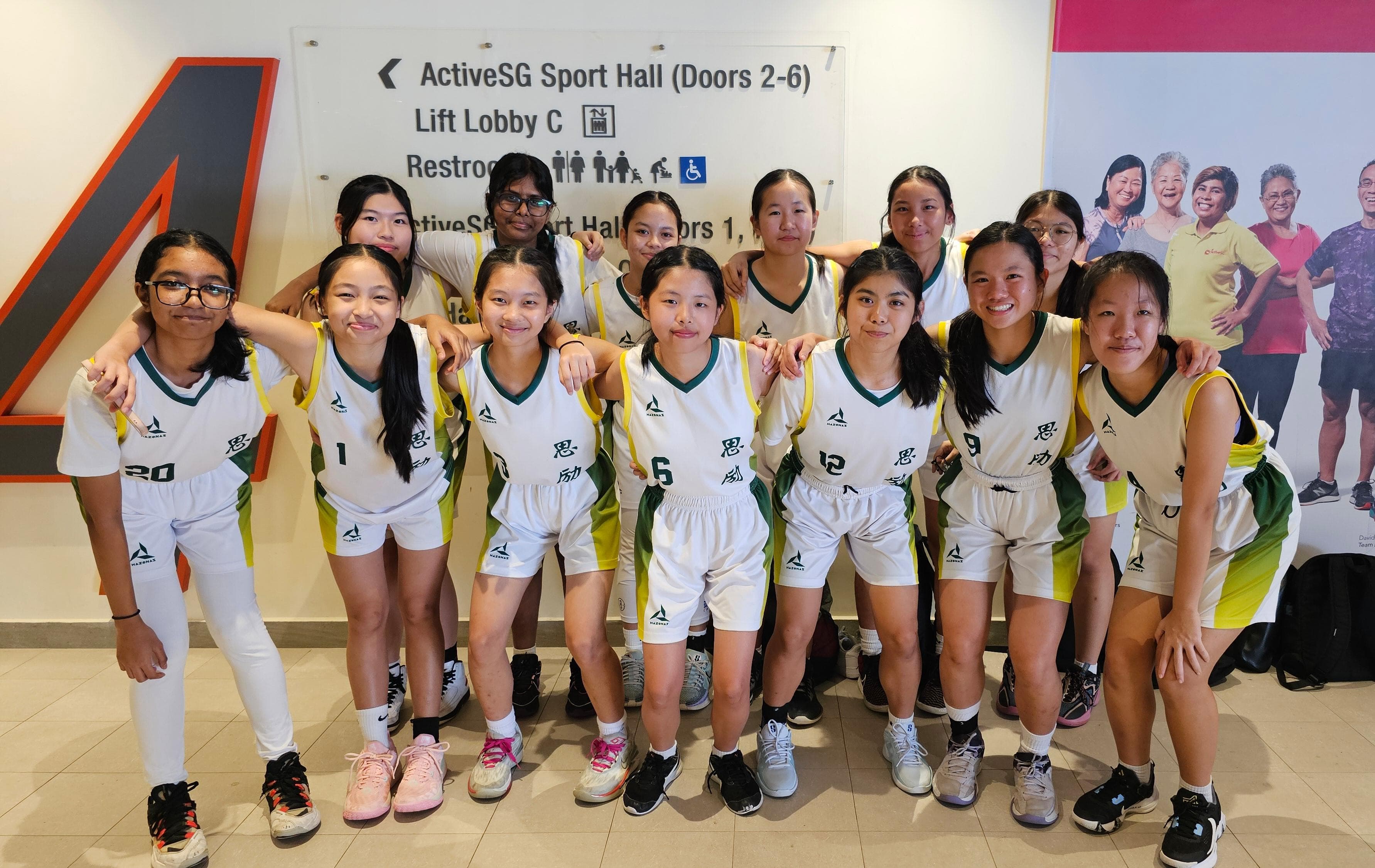 A girls basketball team in white and green uniforms poses together inside an ActiveSG Sport Hall.