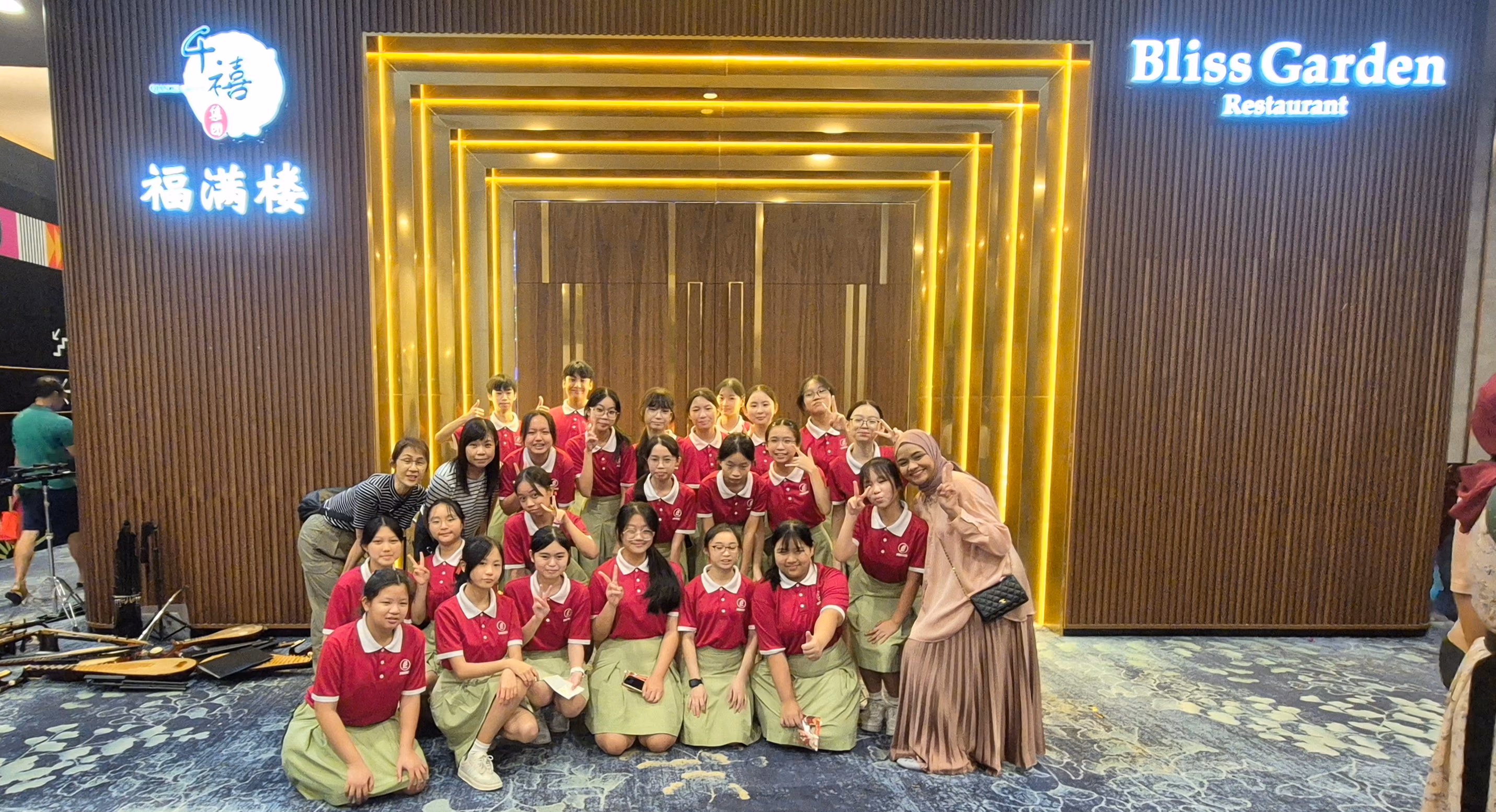 Group of students in red polo shirts and green skirts posing in front of Bliss Garden Restaurant entrance.