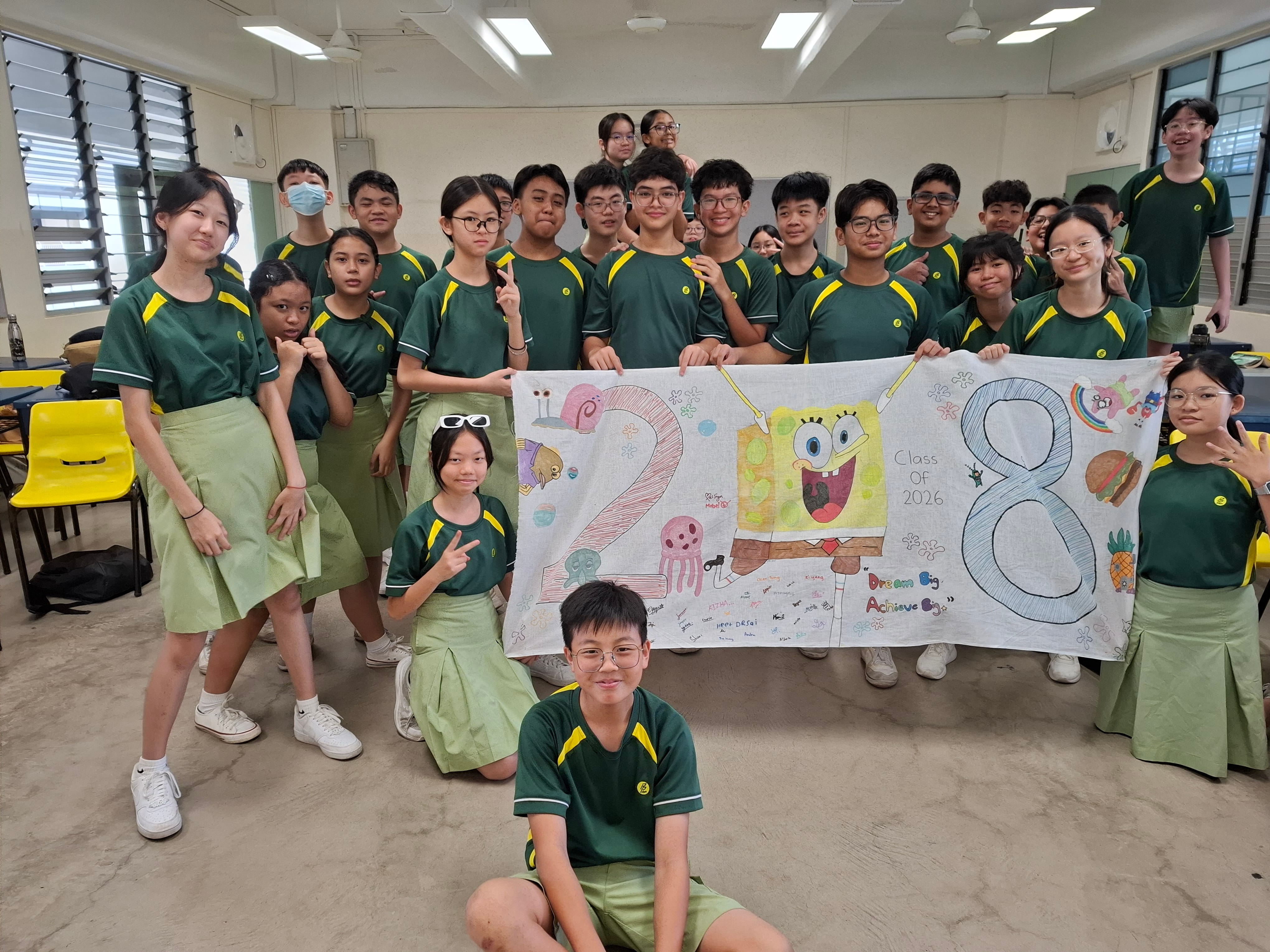 Photograph of a group of students in green and yellow uniforms posing indoors while holding a colorful banner featuring SpongeBob SquarePants, the numbers "2" and "8," and various sea-themed drawings. The banner includes "Class of 2024" text, indicating a celebratory or graduation event for the class.