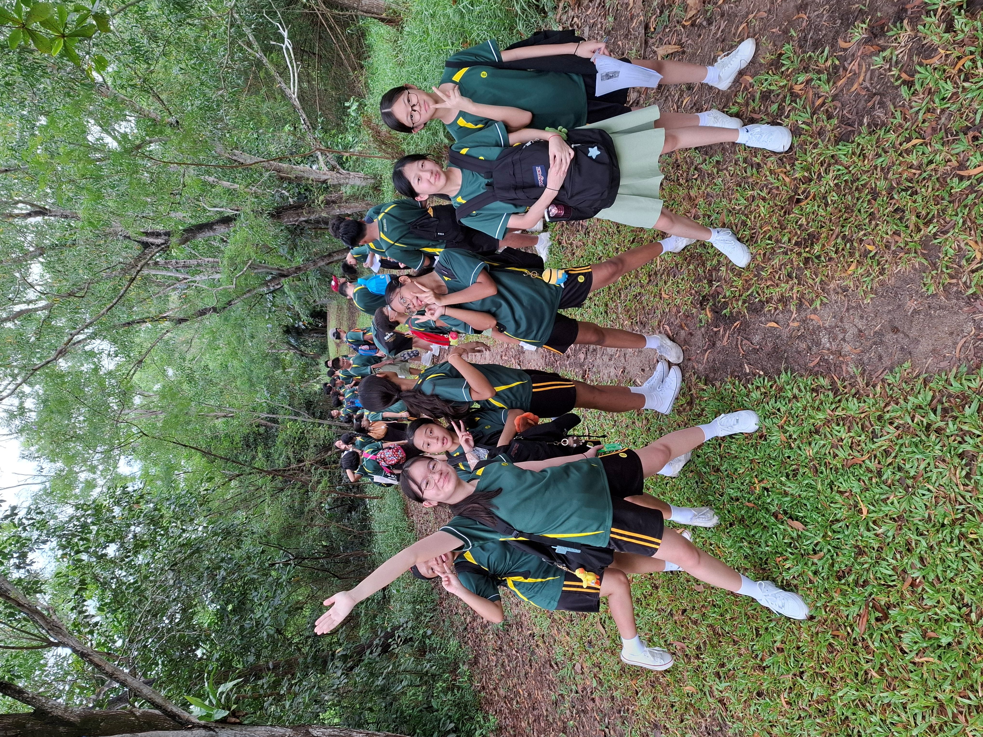 Photograph of a group of students wearing matching green and yellow sports uniforms standing on a grassy path surrounded by trees, likely during an outdoor school event or field trip. Several students in the foreground are smiling and posing with peace signs, while others form a line extending into the background.