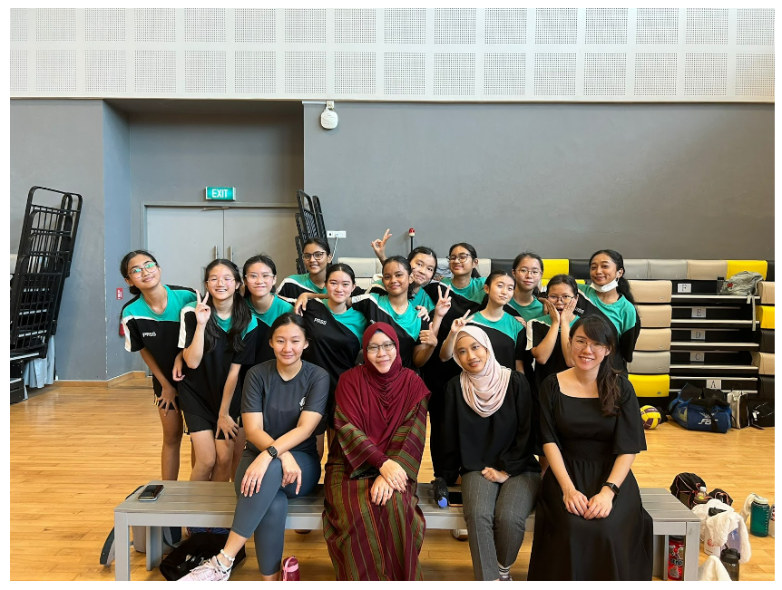 A group of girls in teal and black sports uniforms pose with four adults in a gym, many making peace signs.