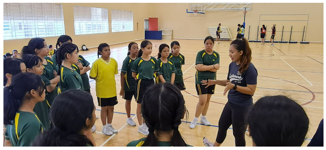 A coach in a navy shirt addresses a group of girls in green uniforms gathered in an indoor sports hall.