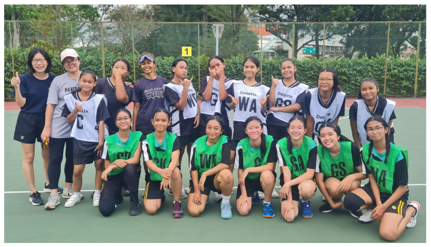 Two netball teams of young girls in green and white bibs pose together on an outdoor court.