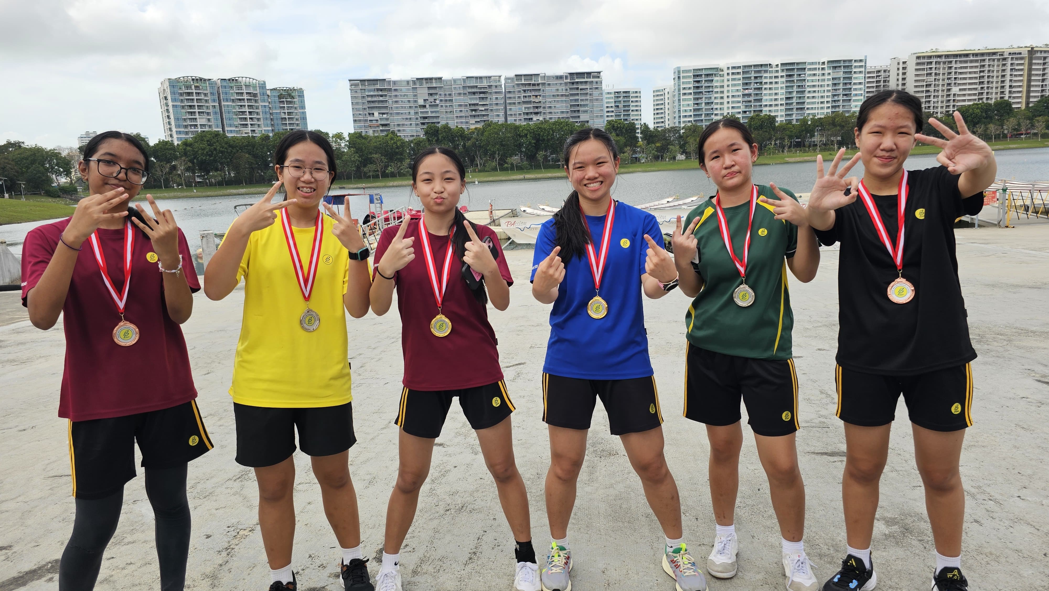 Six young girls wearing medals pose outdoors near a waterway with apartment buildings in the background.