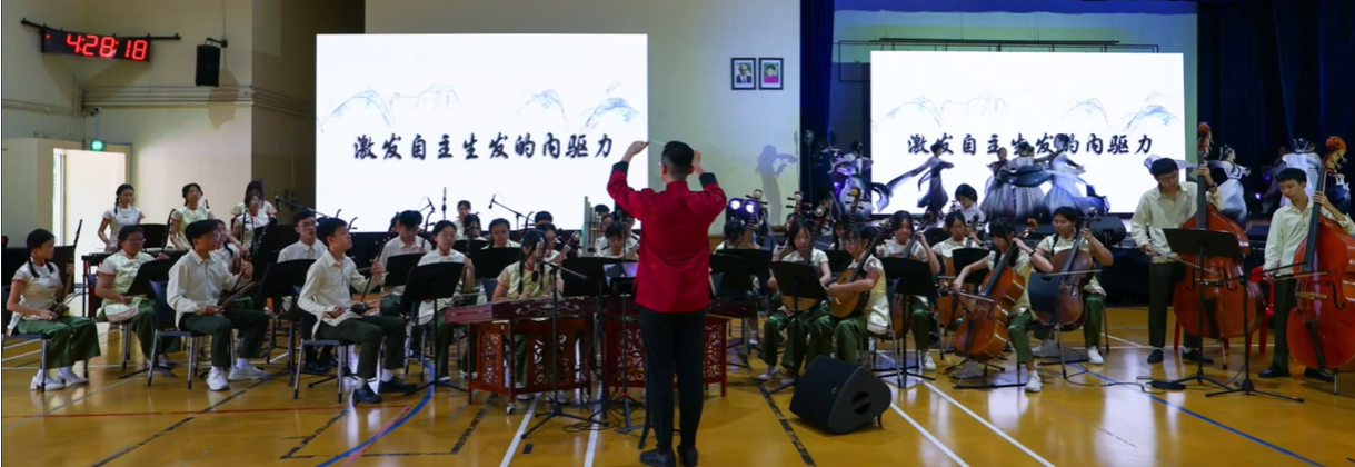 A conductor in red leads a large student orchestra on a gym stage with two Chinese text projection screens behind them.