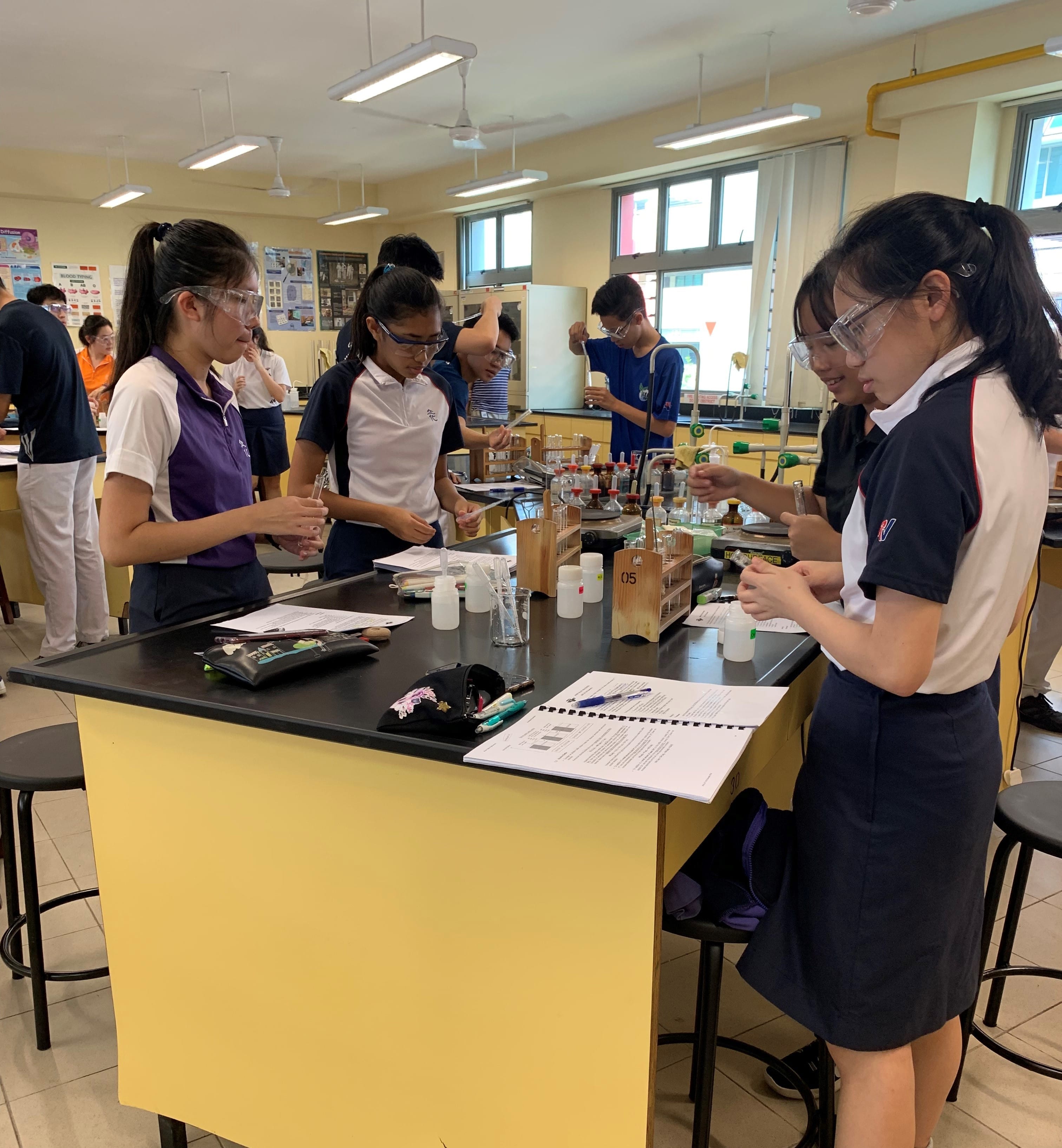 Students in uniforms and safety goggles conduct chemistry experiments with test tubes at lab benches.