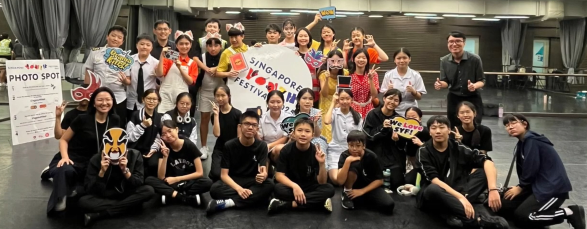 Large group of students posing at a Singapore Youth Festival photo spot, holding props and signs indoors.