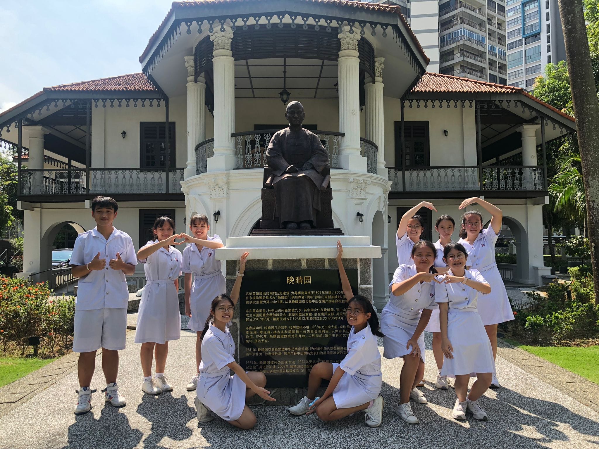 Students in white uniforms pose in front of a bronze seated statue at a colonial-style heritage building with Chinese signage.