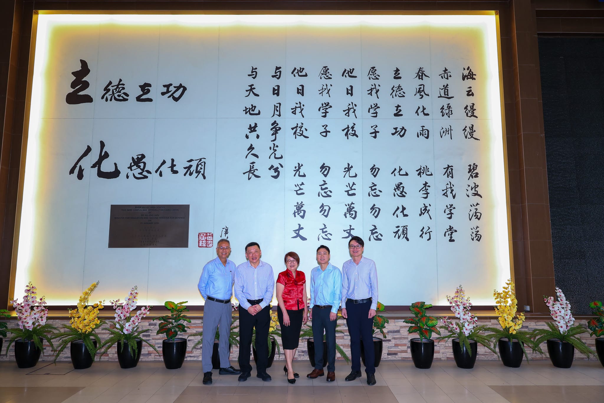 Five people stand posing in front of a large illuminated wall with Chinese calligraphy, flanked by flower arrangements.