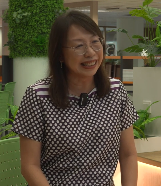 A smiling woman with glasses wearing a black and white polka dot shirt with a lapel mic, seated indoors near plants.