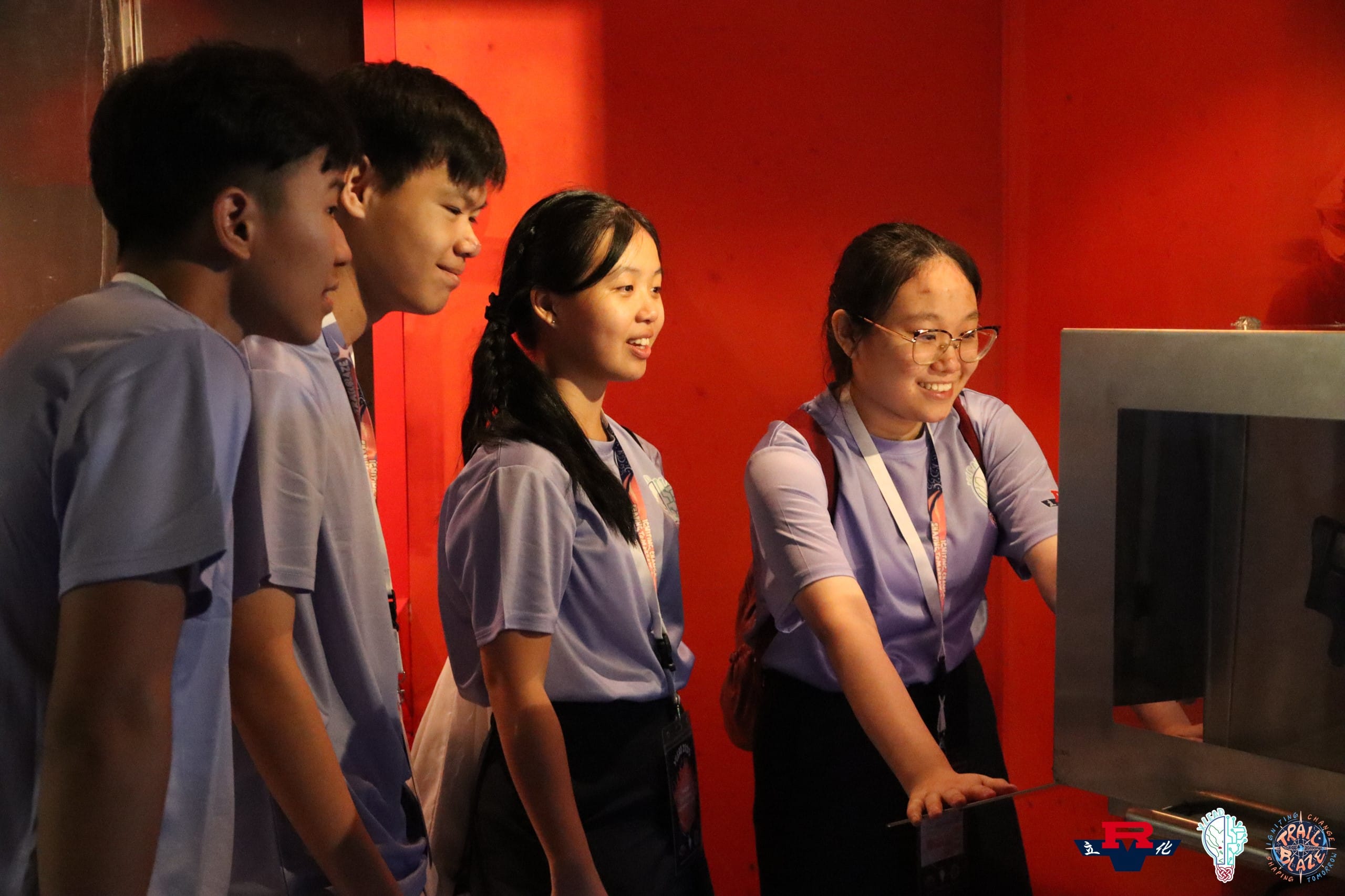 Four young students in blue shirts and lanyards smile while interacting with a display in a red-walled room.