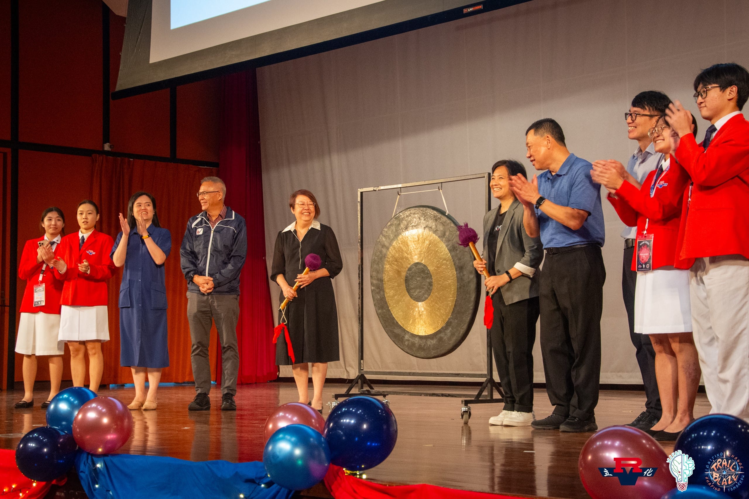 A group of people stand on a stage near a large gold gong, clapping, with balloons in the foreground.