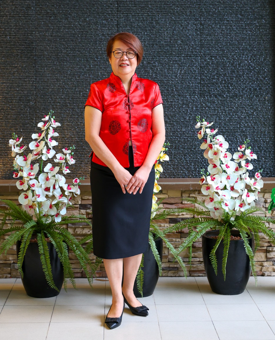 Woman in red silk top and black skirt stands smiling between potted orchids and ferns near a stone wall.