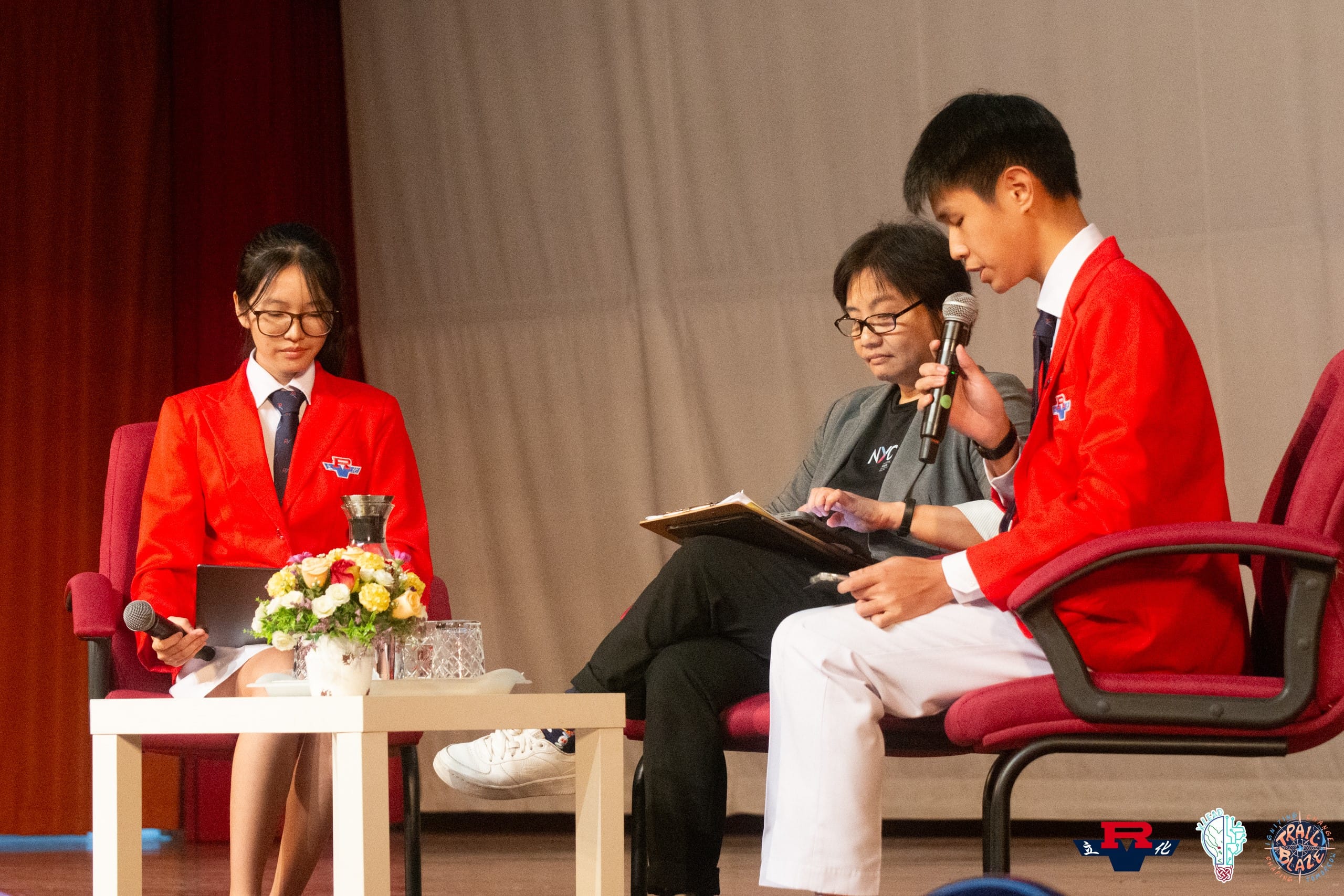 Three people sit on stage; two students in red blazers hold microphones while an adult reviews notes between them.