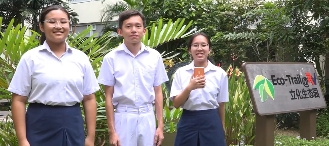 Three students in white uniforms stand outdoors beside an Eco-Trail sign with Chinese characters and green plants behind them.