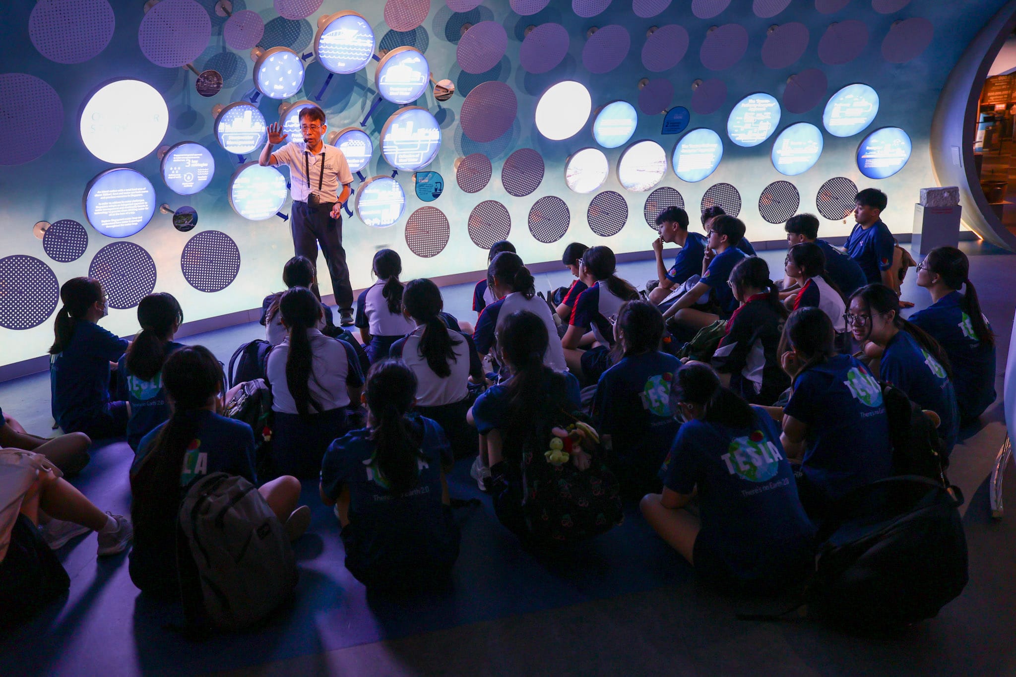 A man presents to students seated on the floor in a museum room with glowing circular displays on the walls.