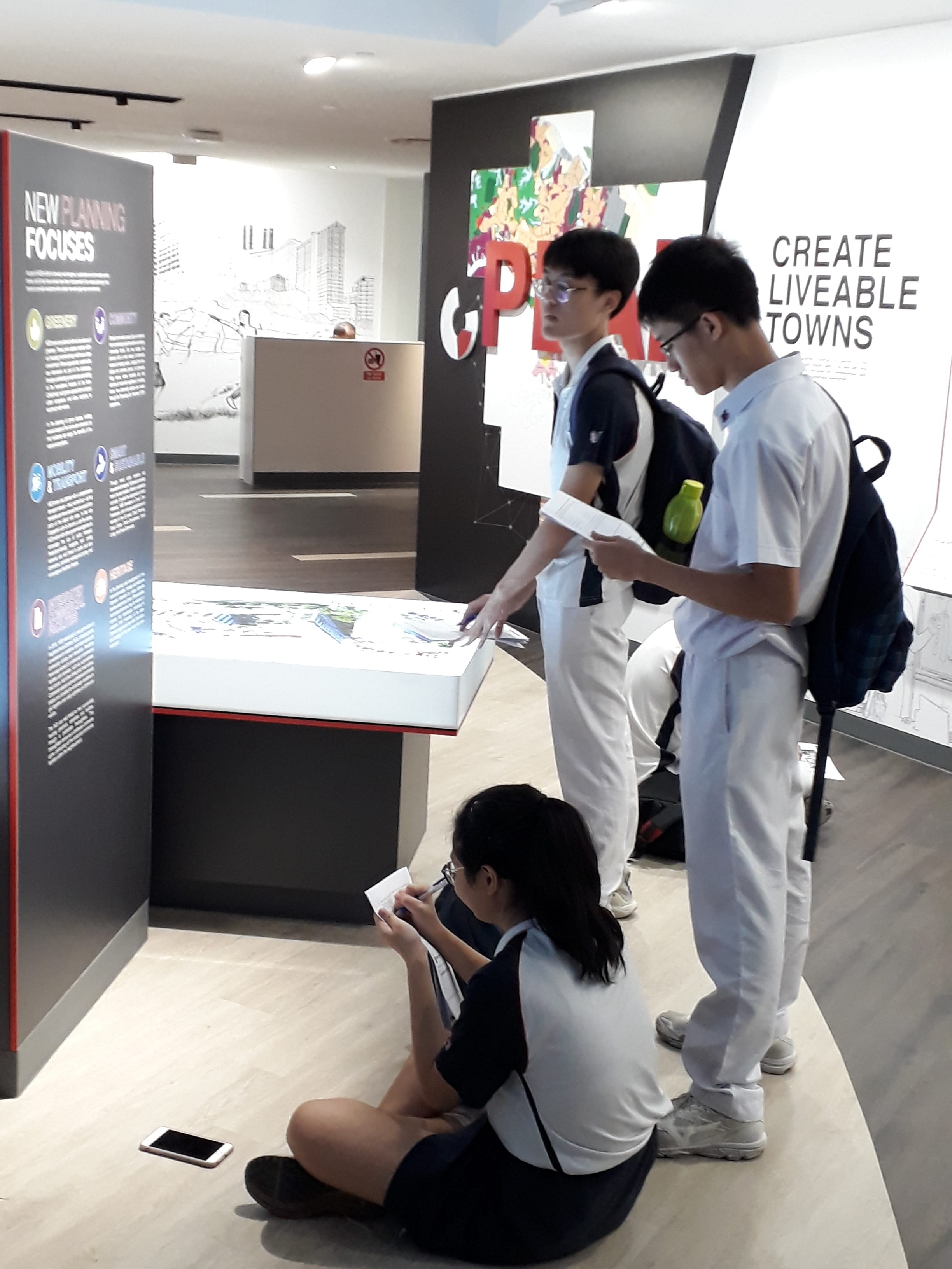 Three uniformed students examine exhibits at a urban planning display with a "Create Liveable Towns" wall sign.