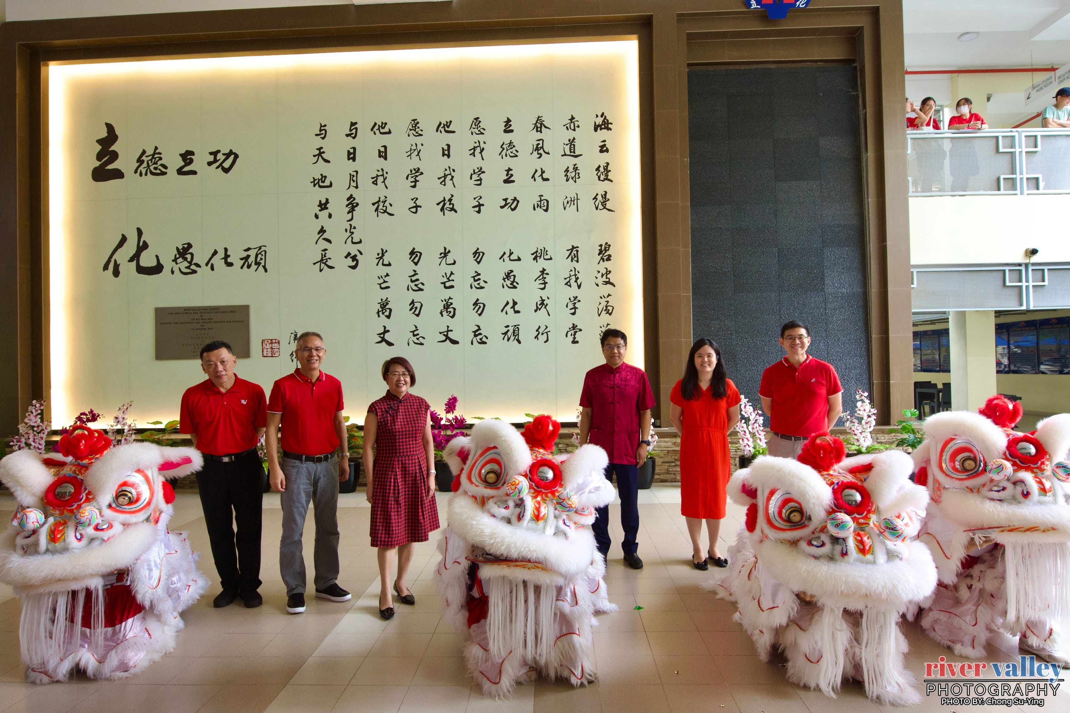 Five people in red attire stand before a Chinese calligraphy wall, flanked by three white and red traditional lion dance costumes.